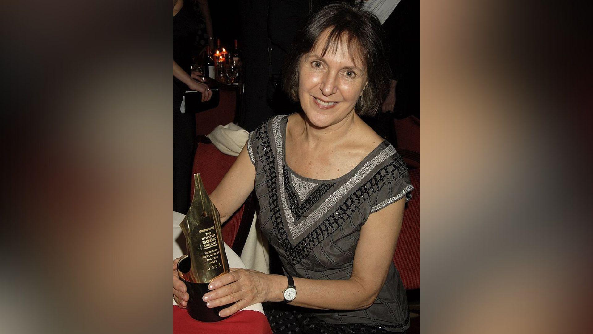 A woman seated at a table holding an award shaped like the nib of an ink pen.  She has dark hair and brown eyes and is smiling at the camera.  She is wearing a grey top embellished with silver and black shiny beads