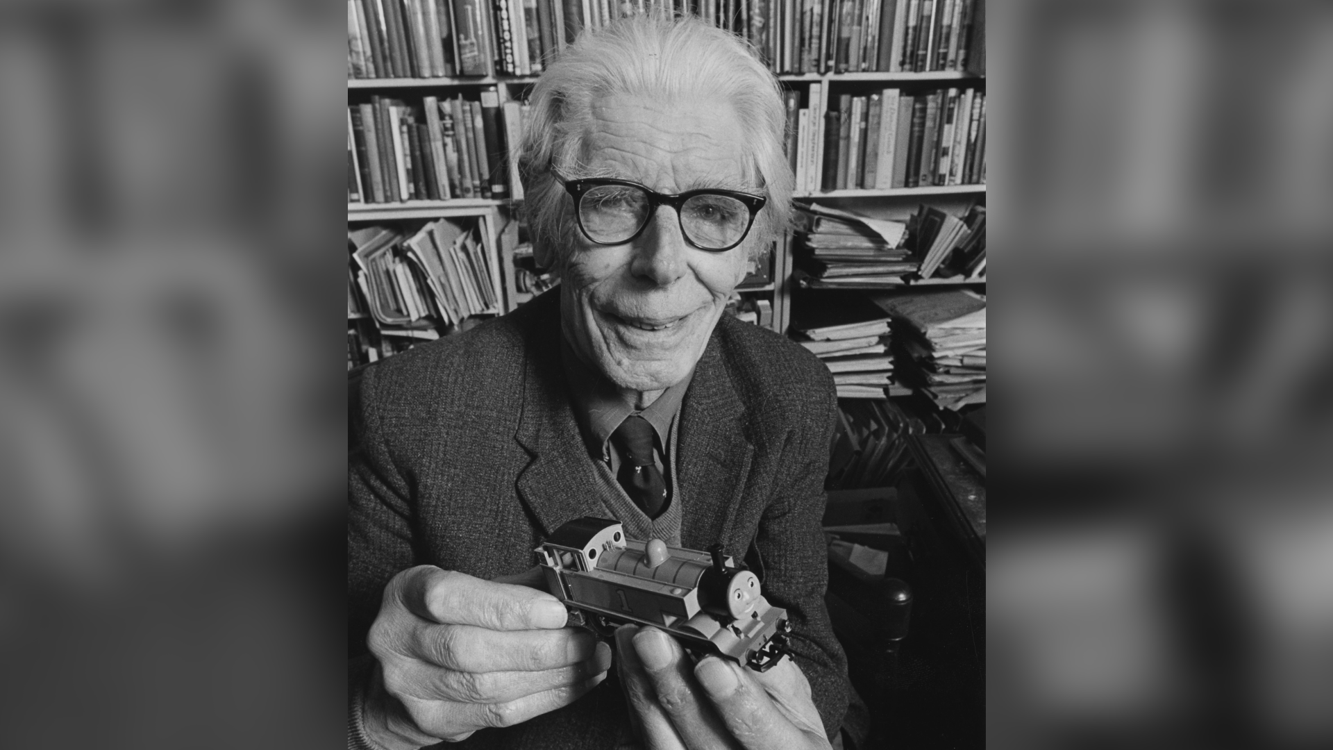 A black and white photo of Reverend Wilbert Awdry. He is an elderly man with fair, short hair. He is wearing a shirt, tie and a blazer. He is holding a small model of Thomas the Tank Engine, a children's steam train character. It has a smiley face.