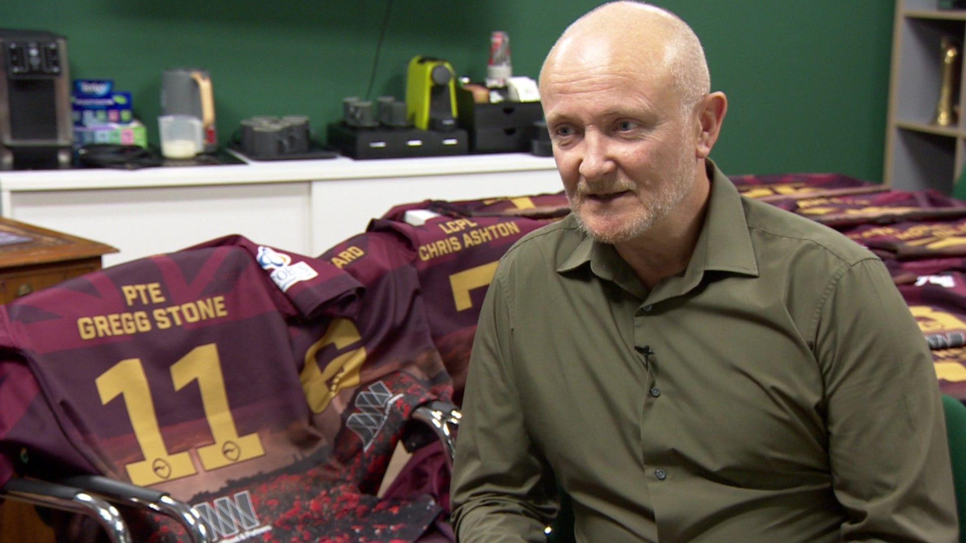 Jim Fishburn is sitting in front of the maroon rugby shirts which are laid out on a table behind him. The names and numbers on the shirts are in yellow.  The shirt immediately behind Mr Fishburn is that of PTE Gregg Stone, who was killed in Afghanistan in 2012.