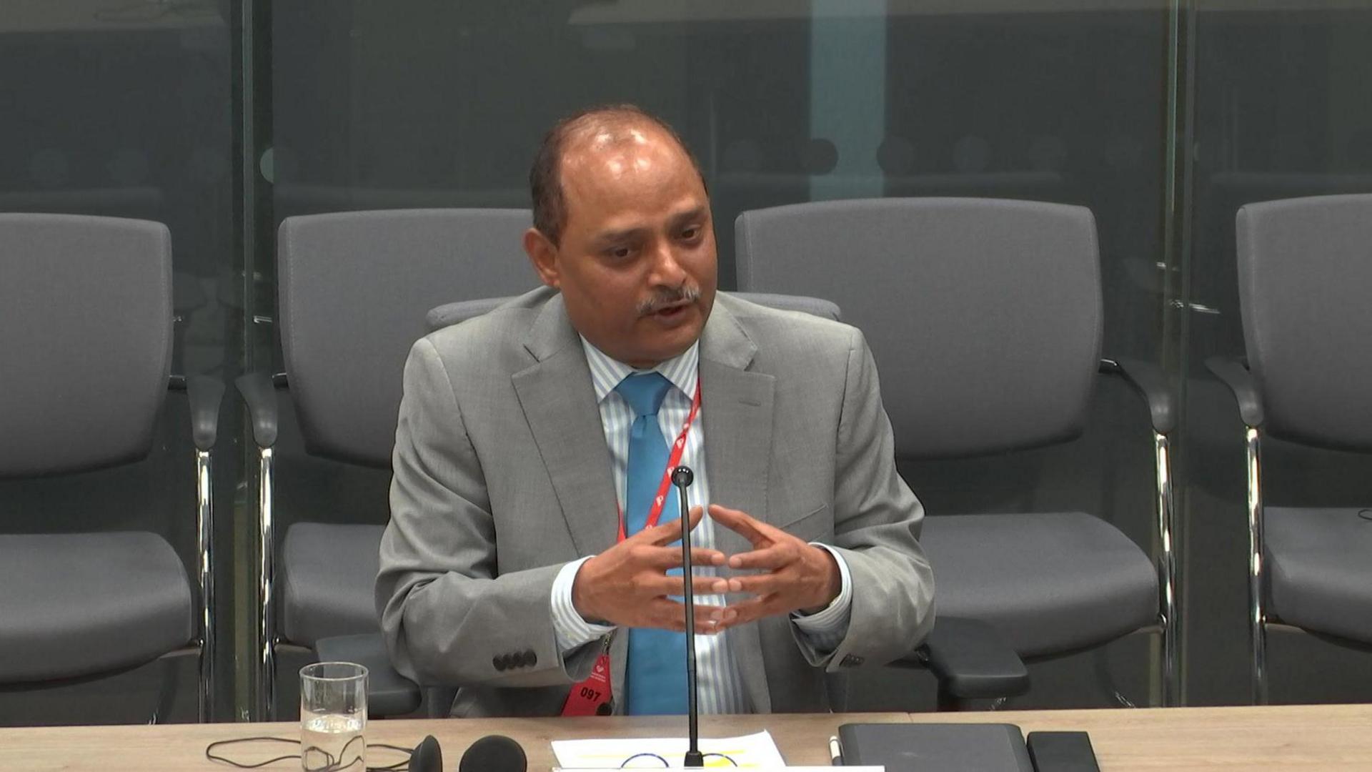 Rajesh Nair gesturing with his hands as he speaks to Senedd members in the economy committee. He is sitting behind a desk with a row of chairs behind him and is wearing a light grey suit, striped grey and white shirt and a light blue tie.