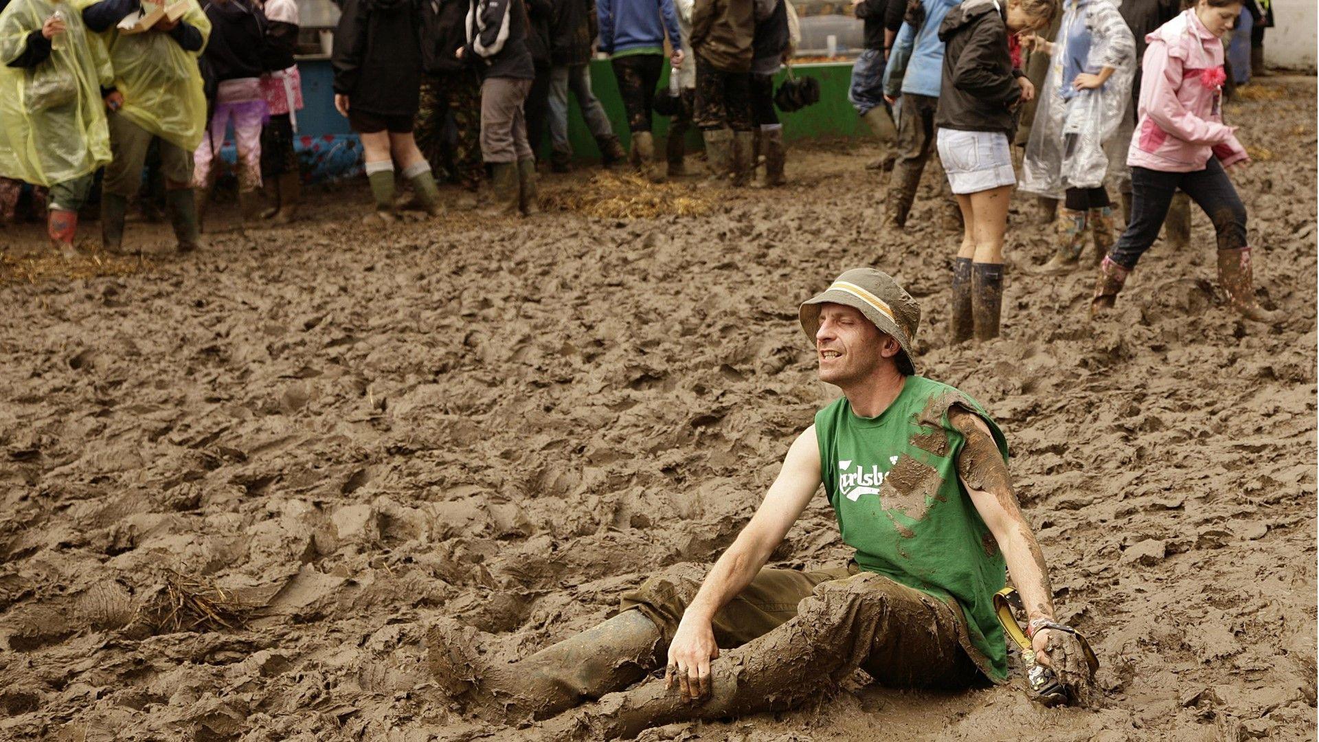 A muddy walkway at Glastonbury with festival-goers wearing waterproofs and wellies. One man in the foreground is very muddy and sitting in the mud