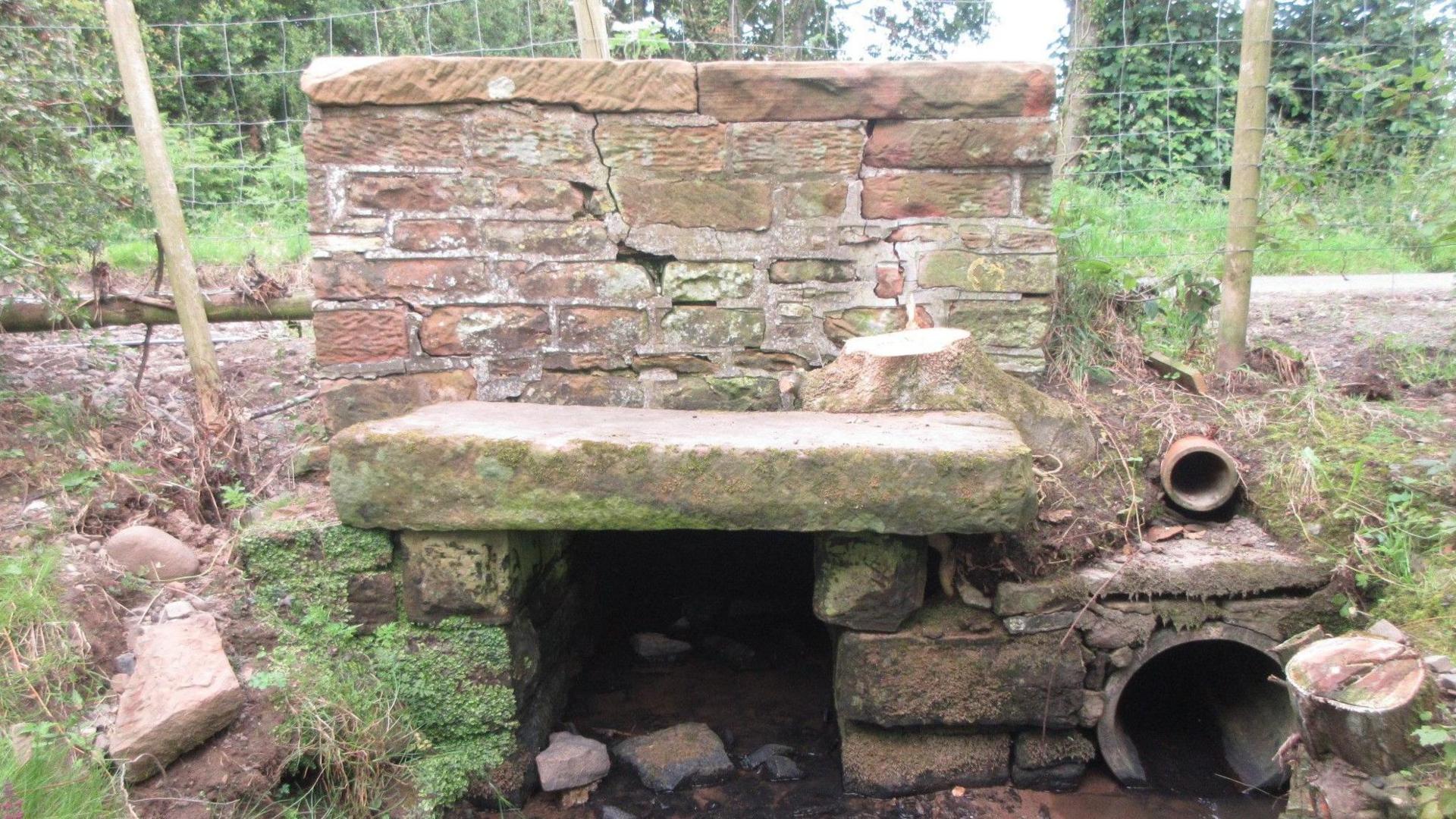 An ageing brick and stone culvert spanning a fast-flowing stream. On the muddy banks to either side are leafless trees, with more in the background alongside a narrow road.
