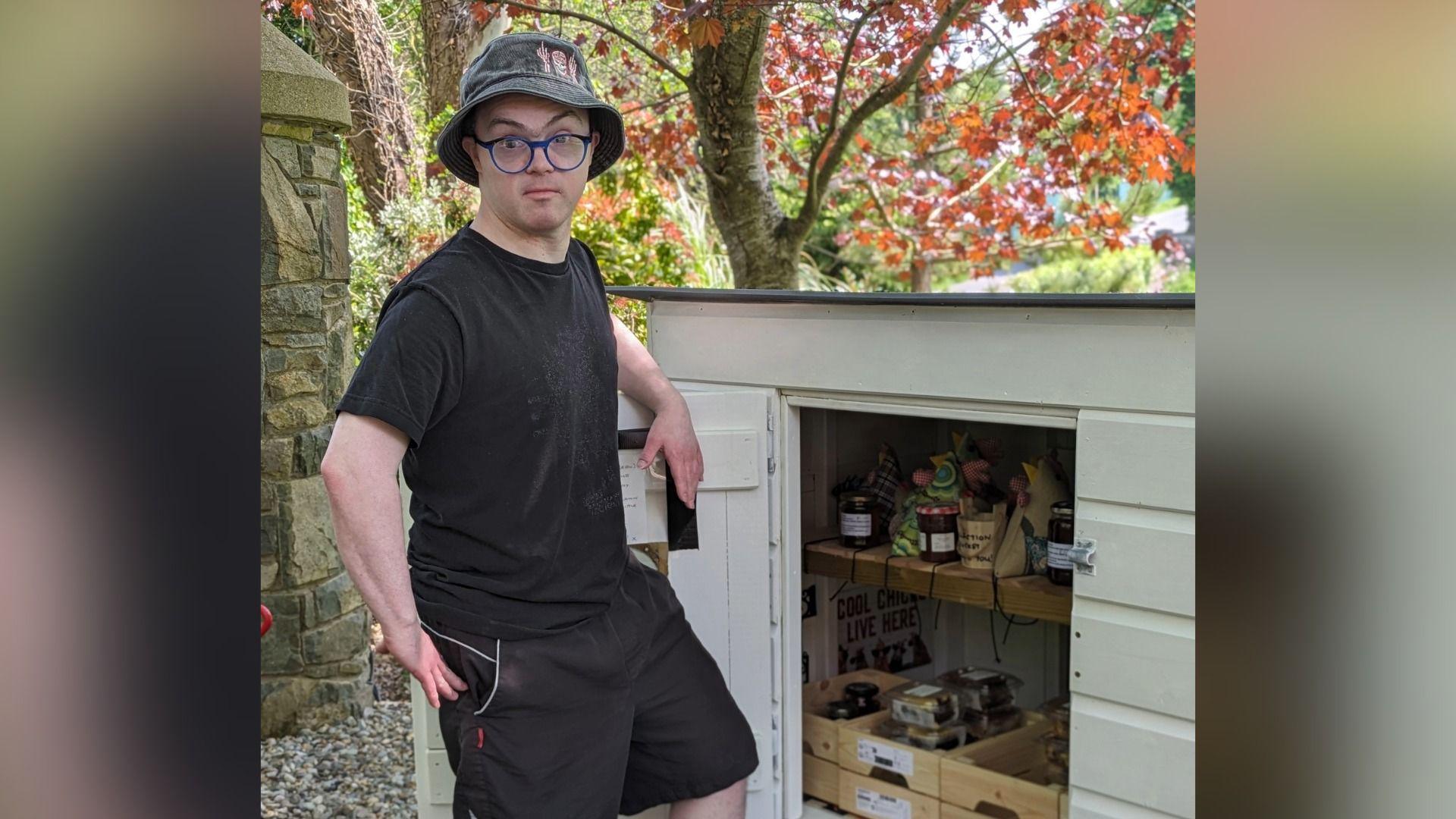 Young man smiling at camera. Wearing black t-shirt, black hat, glasses and shorts. The honesty box is pictured beside him. It is a cream white colour and has wooden baskets inside containing produce to sell. The picture is outdoors. 