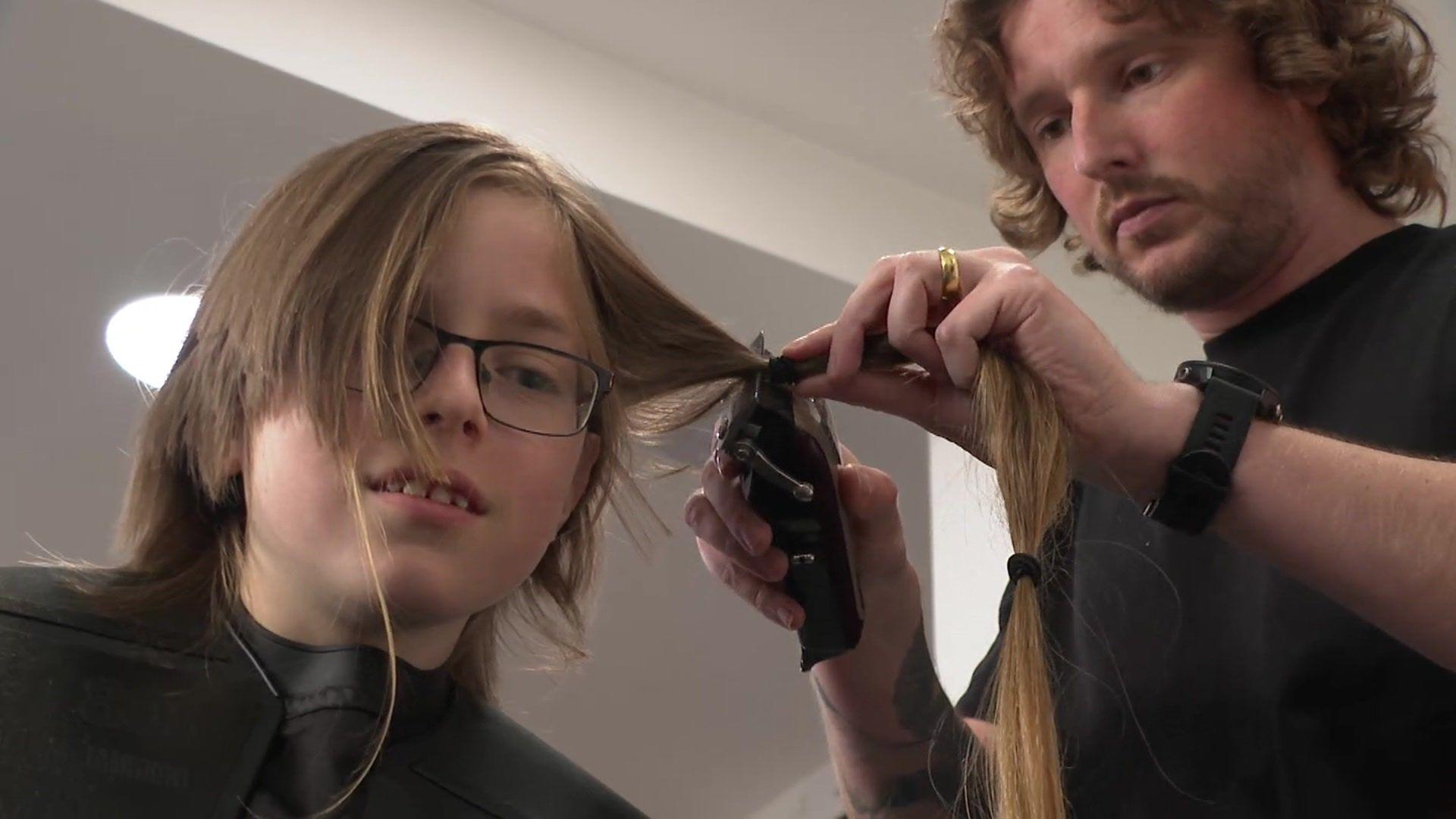 A young boy in glasses sits having his hair cut by a man holding a pair of hair clippers