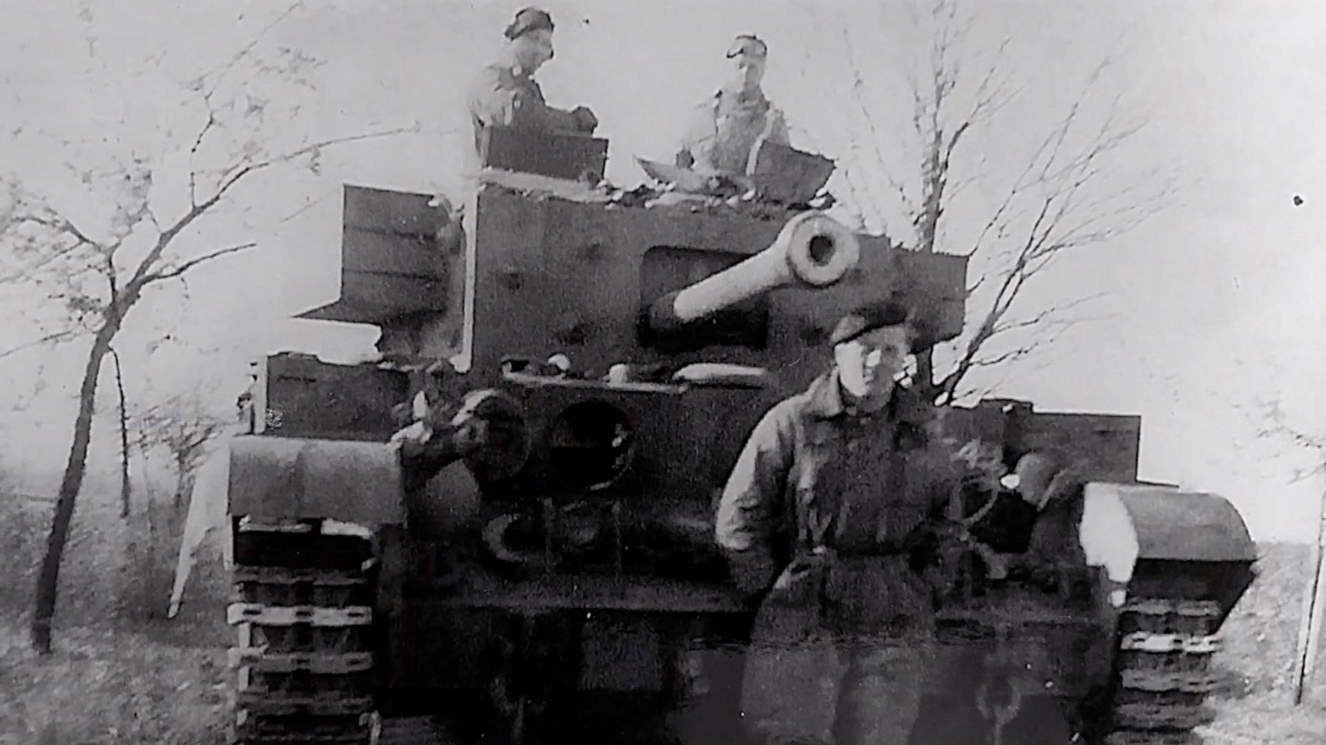 A black and white photo showing Richard Aldred standing in army uniform in front of a tank. On the tank are two other men also wearing army uniform. There are a few slim trees behind them.