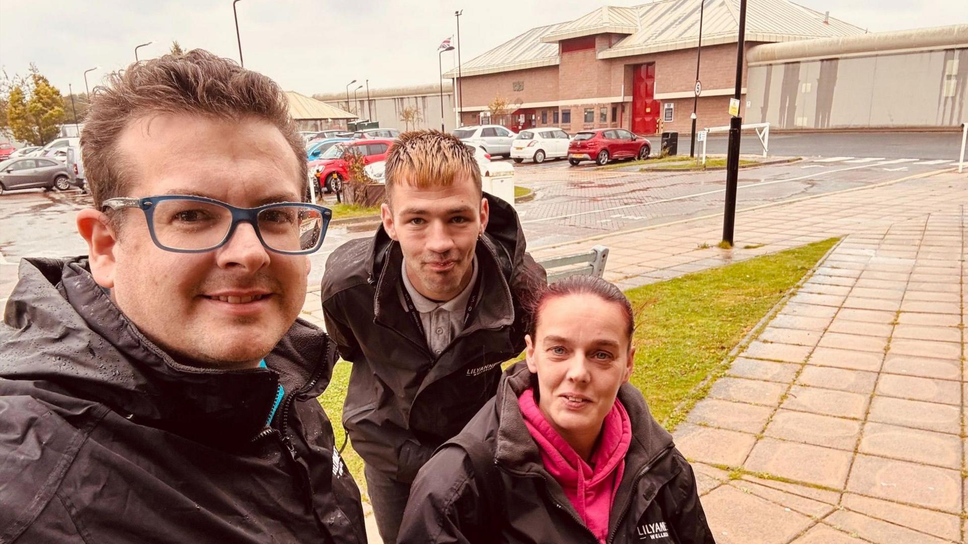 Trevor Sherwood in black jacket and glasses. He is standing next to Kieron Higgins who is smiling, as well as Angela Arnold who is wearing a pink hoodie and a black jacket. They are in a car park. 