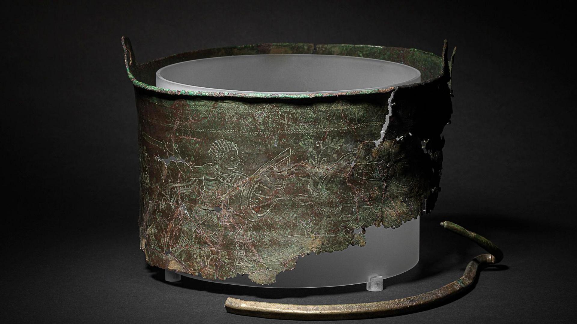 A photo of the Bromeswell Bucket which has been displayed on a black surface with a black background. Part of the bucket is missing and etchings can be seen on the bucket's surface.