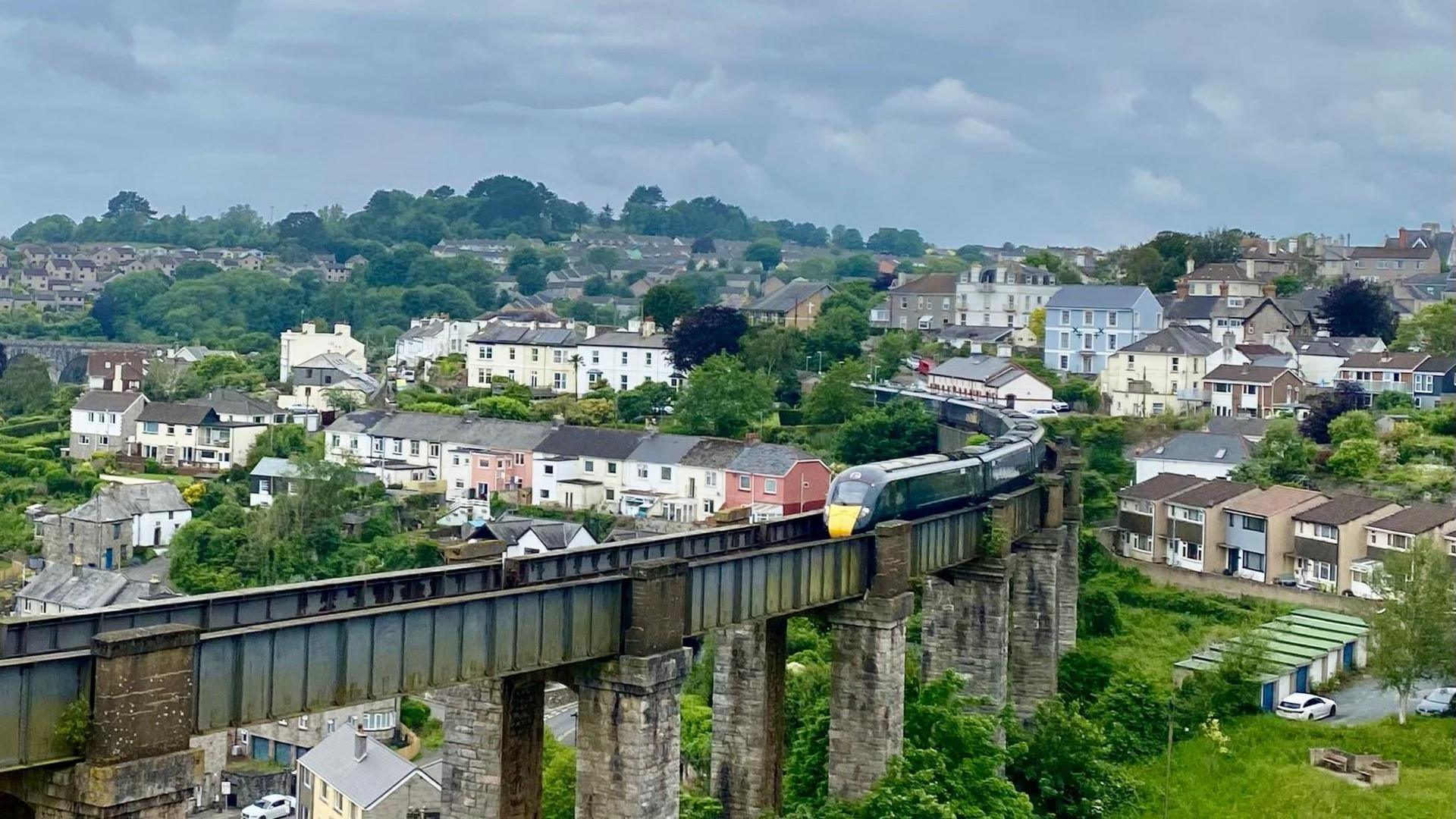 A green train with several carriages travels along a bridge above Saltash in Cornwall on an overcast day.