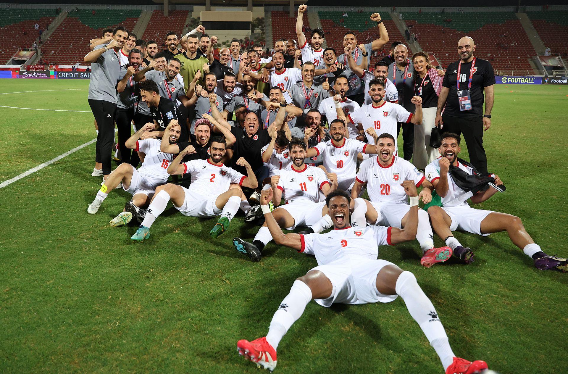 A group picture of Jordanian football players and other supporting members of the wider team smiling and celebrating on a football pitch.  Many of the players are wearing white kit and sitting on the ground. They are all looking at the camera.