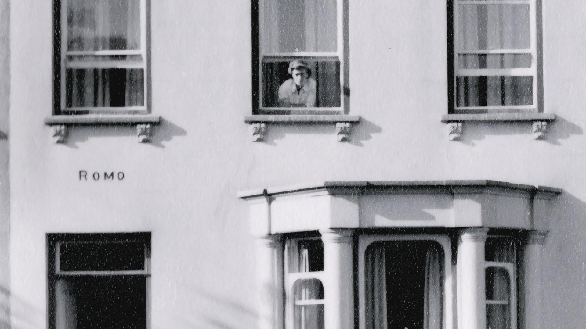 A black and white picture of the front of a building. There are three sash windows open on the first floor and a lady leans out of the middle one. Below is a bay window with curtains and above the doorway on the lefthand side is the name of the house - Romo.