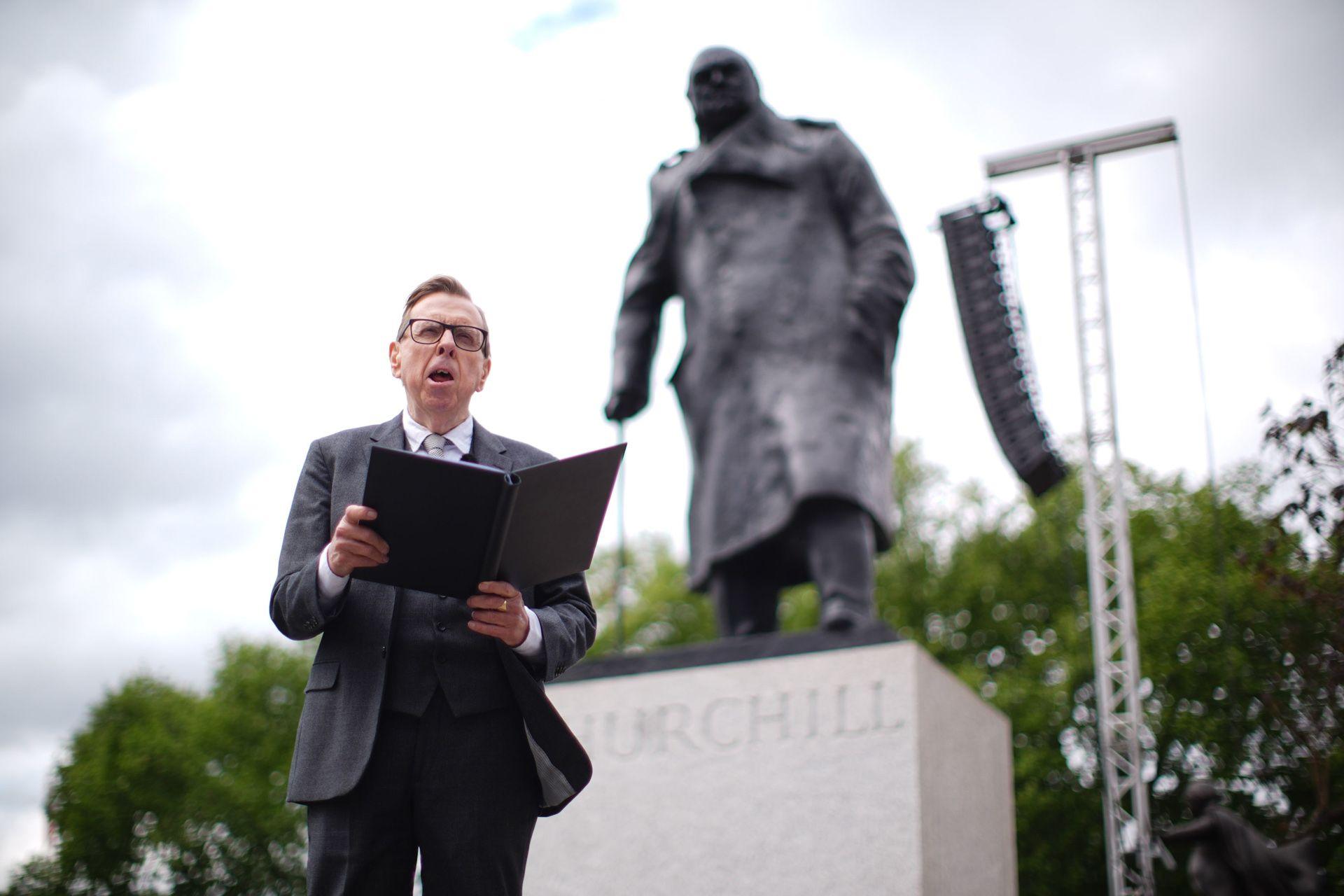 Actor Timothy Spall wears a suit and tie as he reads from an A4 booklet in front of the statue of Churchill in Parliament Square.