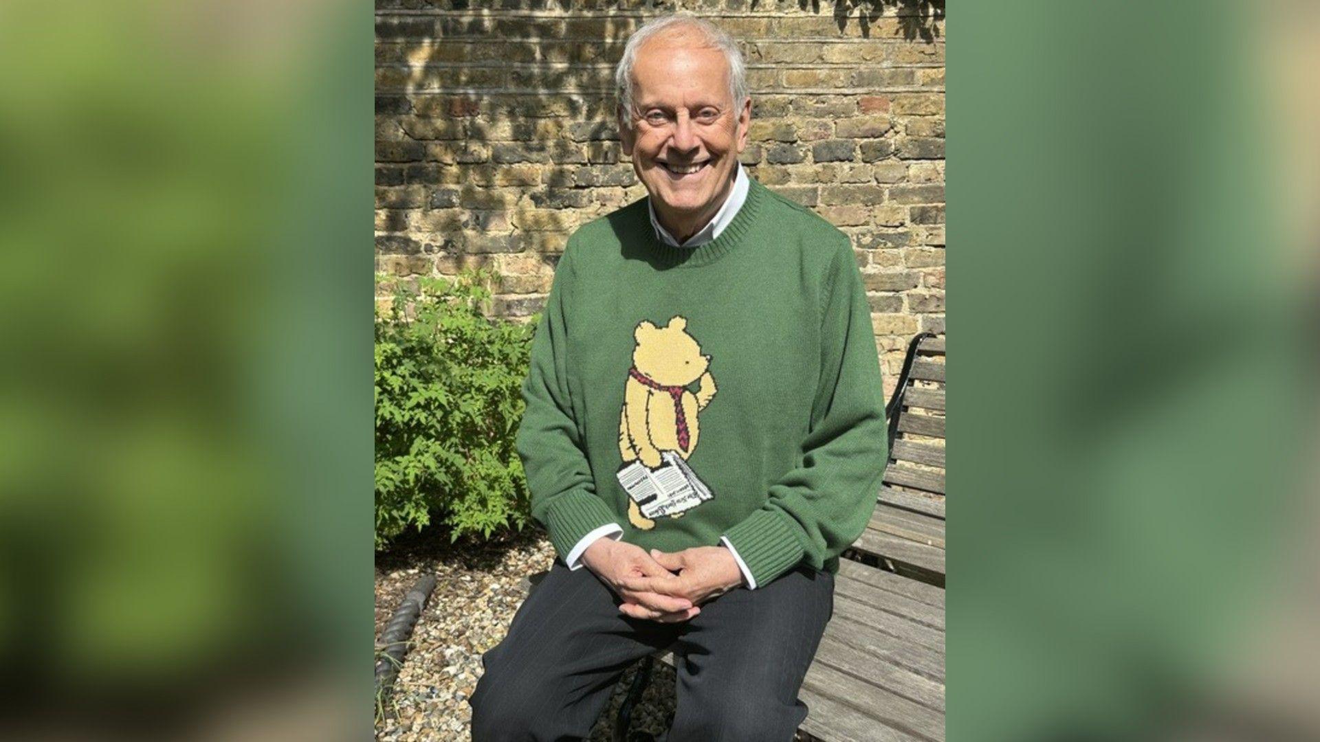 Gyles Brandreth sat on a bench smiling and wearing a green knitted Winnie the Pooh jumper and dark trousers 