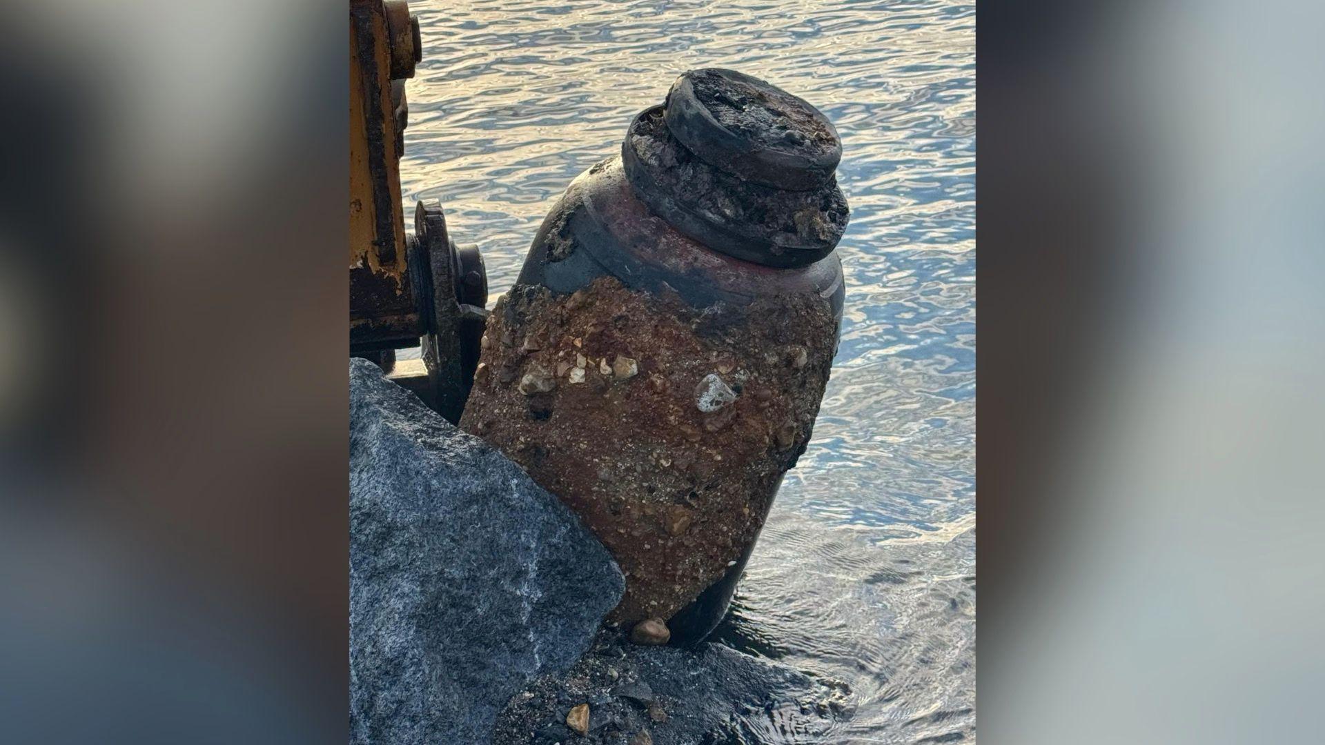 A digger lifts the ordnance out of the water. Close up shot of the weapon, with a yellow digger clasp around it. It is a large, dark grey colour with signs of erosion and rusty colour rocks stuck to the right hand side of it.