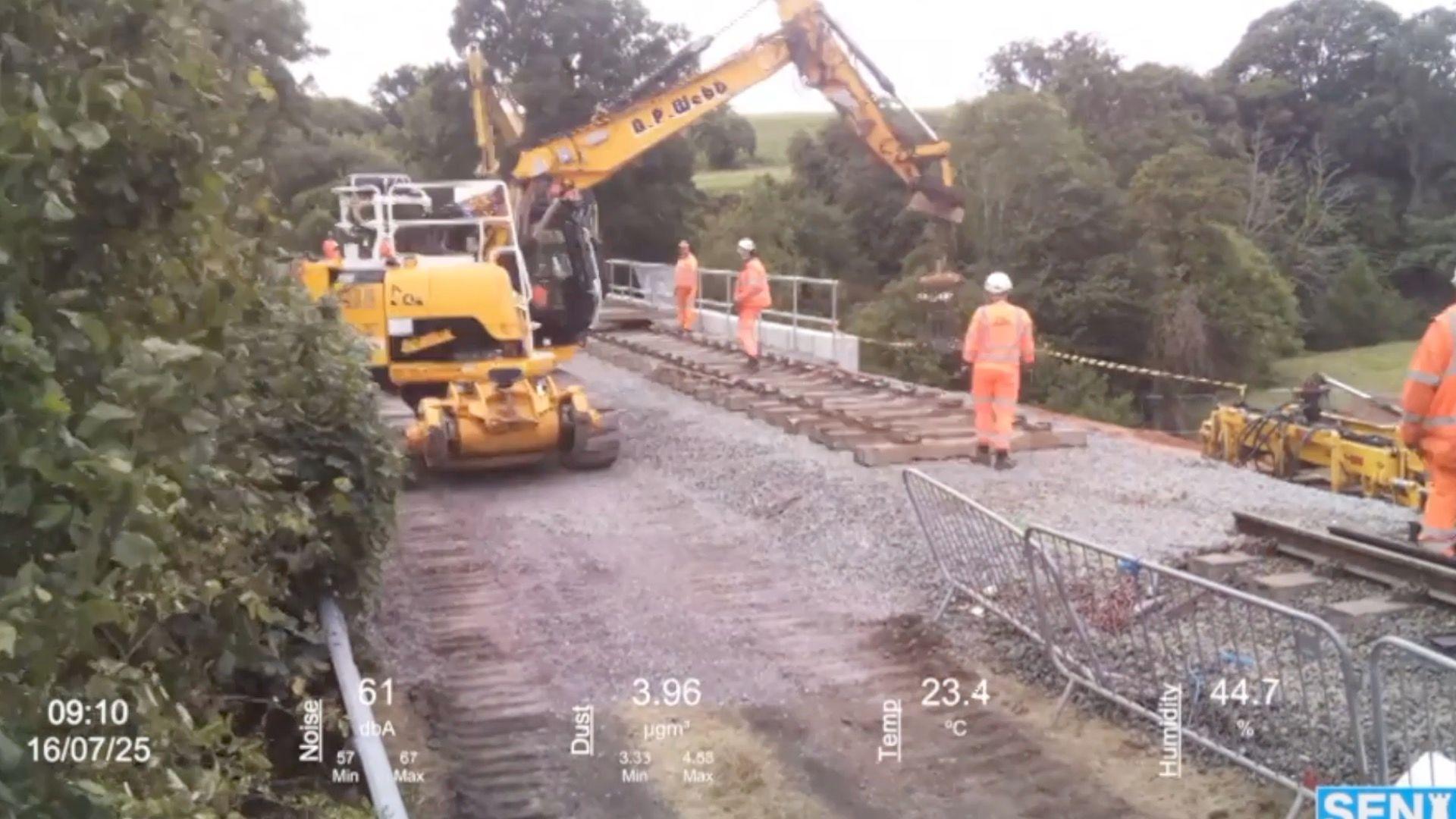 A yellow digger lowers new track on to a railway line. There are four engineers in orange uniforms supervising the work