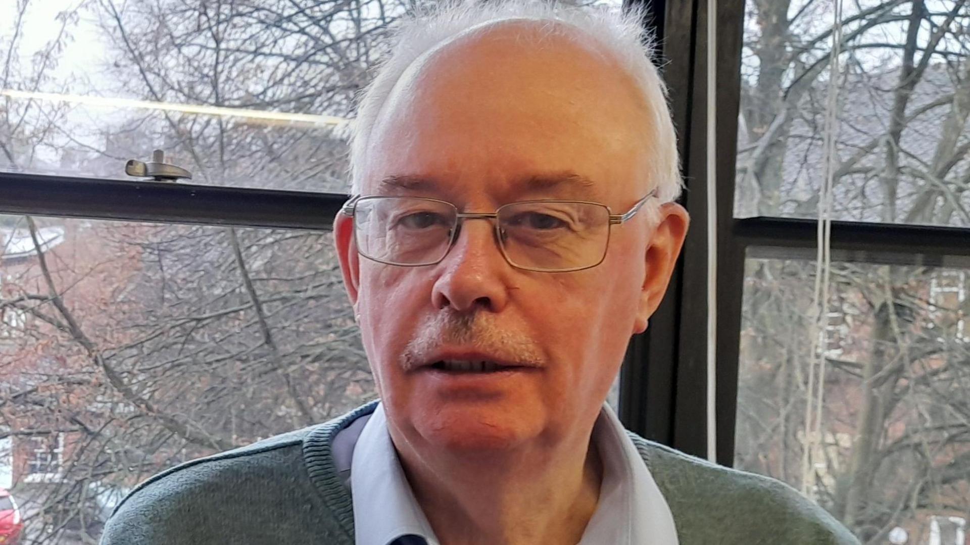 Donald has short receding grey hair and a stubbly-moustache. He is wearing metal-framed rectangle specs on his face, a grey jumper over a blue shirt.