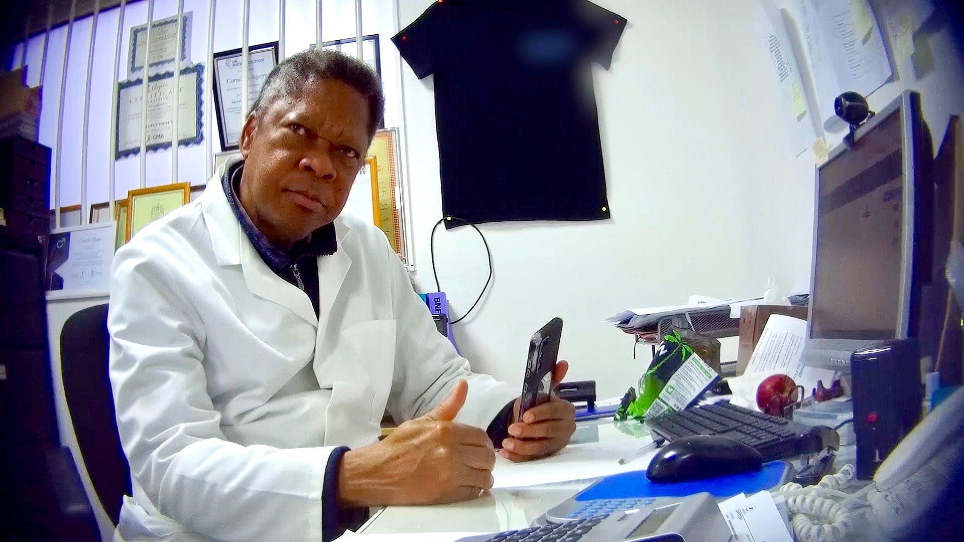 A man in a white lab coat sits at a desk holding a mobile phone. Certificates hang on the wall behind him. He looks directly at the camera inside a pharmacy office