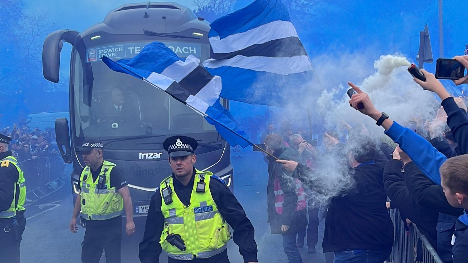 Uniformed police officers walk in front of an Ipswich Football team football bus. Fans wave flags and flares from behind fences. 