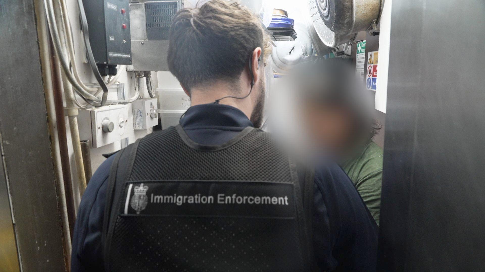 The back head and shoulders of an immigration officer, with a black gilet showing the badge 'immigration enforcement on the back. In the background there is a blurred out photo of someone wearing a dark green top. They are facing each other in a small cupboard area with switches and wires on the left side.