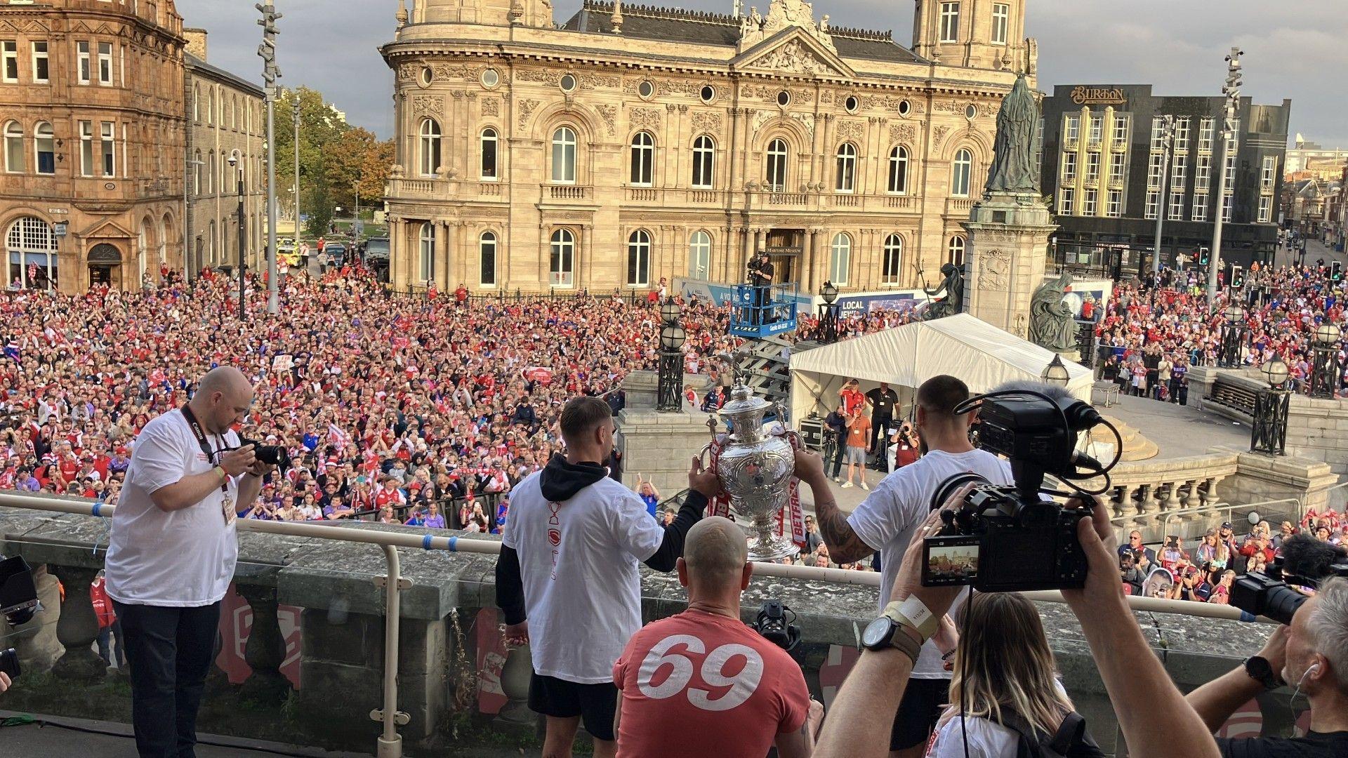 Two players wearing white t-shirts hold aloft a cup on the balcony. There are thousands of fans in front of them.