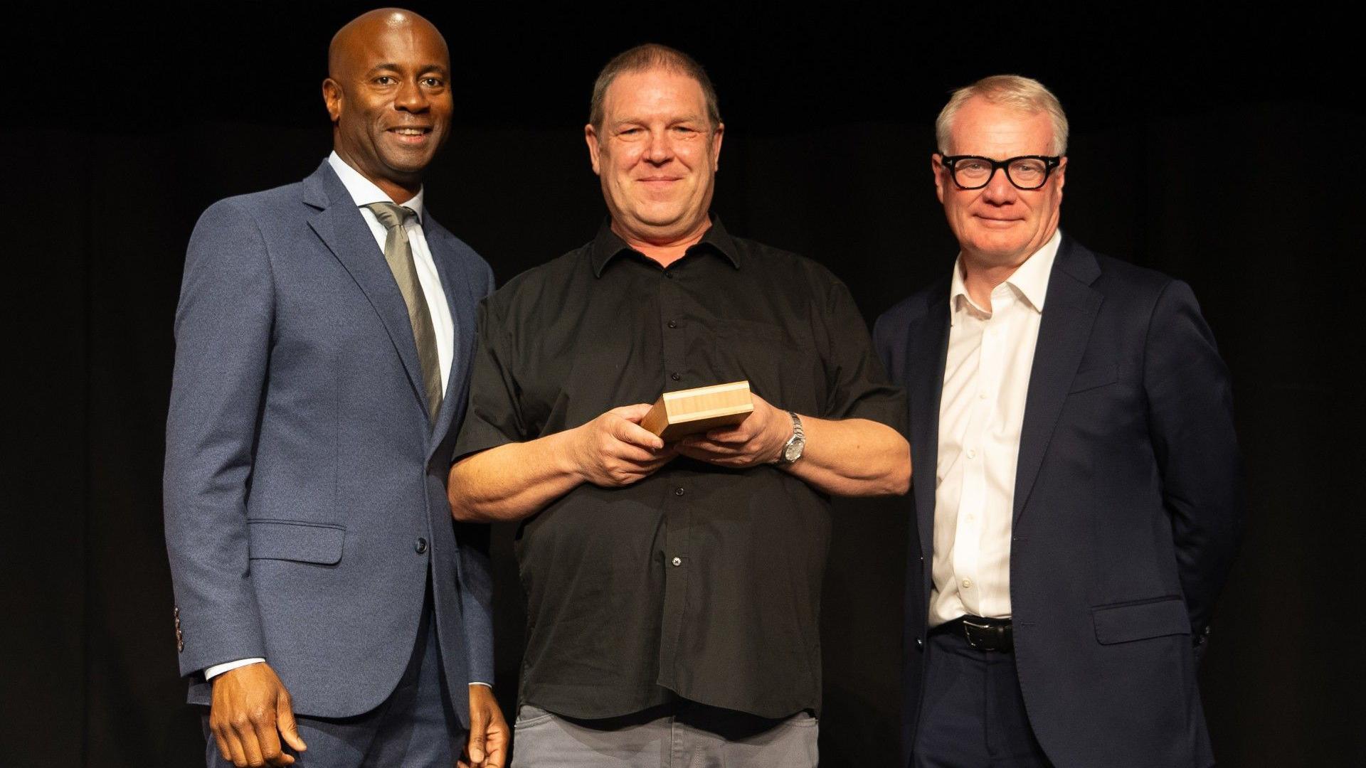 Chris Bruce with actor Nicholas Bailey and West Midlands mayor Richard Parker. Chris has short dark hair and wears a black shirt and grey trousers while holding a box. To his right is Richard Parker in a dark blue suit and white shirt, he wears black glasses. Nicholas Bailey is on the left and has a shaved head and wears a lighter blue suit, a white shirt and olive-green tie.