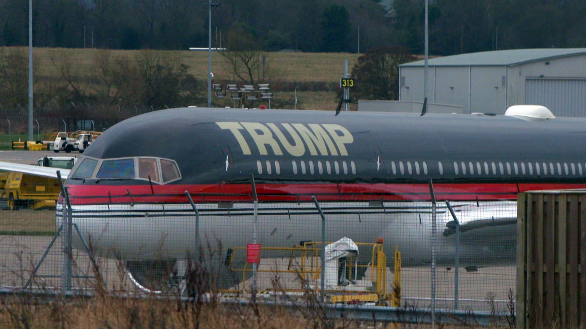 Grey and red plane with TRUMP written on the side in big letters. It is stationary behind a fence at an airport 