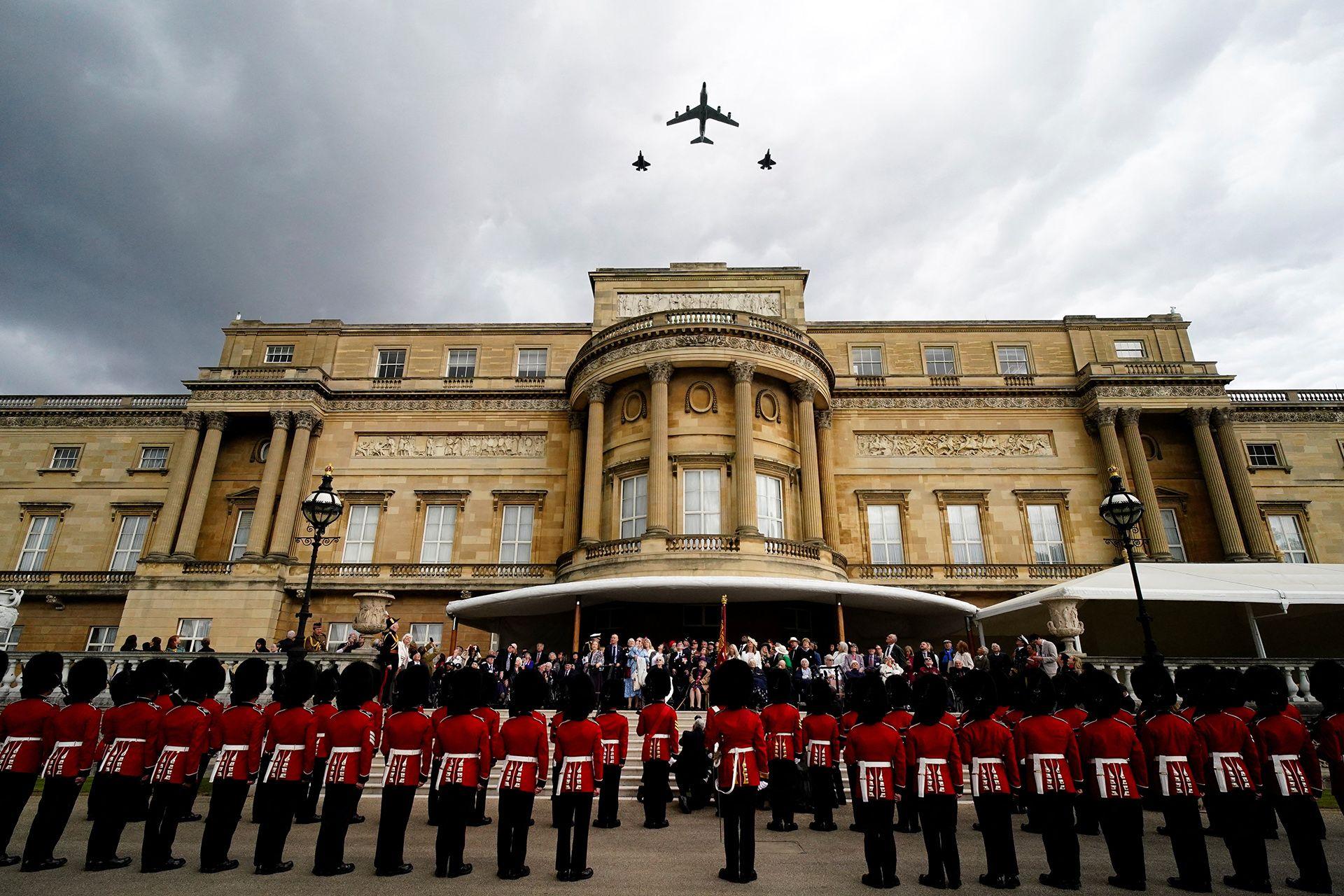 Veterans and members of the armed forces gather outside Buckingham Palace and gaze up to watch RAF aircraft fly over the palace against a cloudy grey sky.