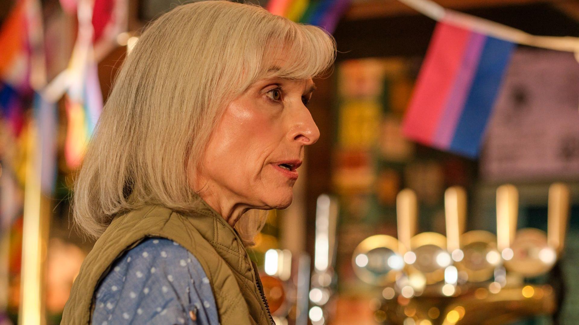 Amelia Bullmore stands in a pub. She has shoulder-length red hair. She is wearing a sleeveless body warmer and a blue top.
