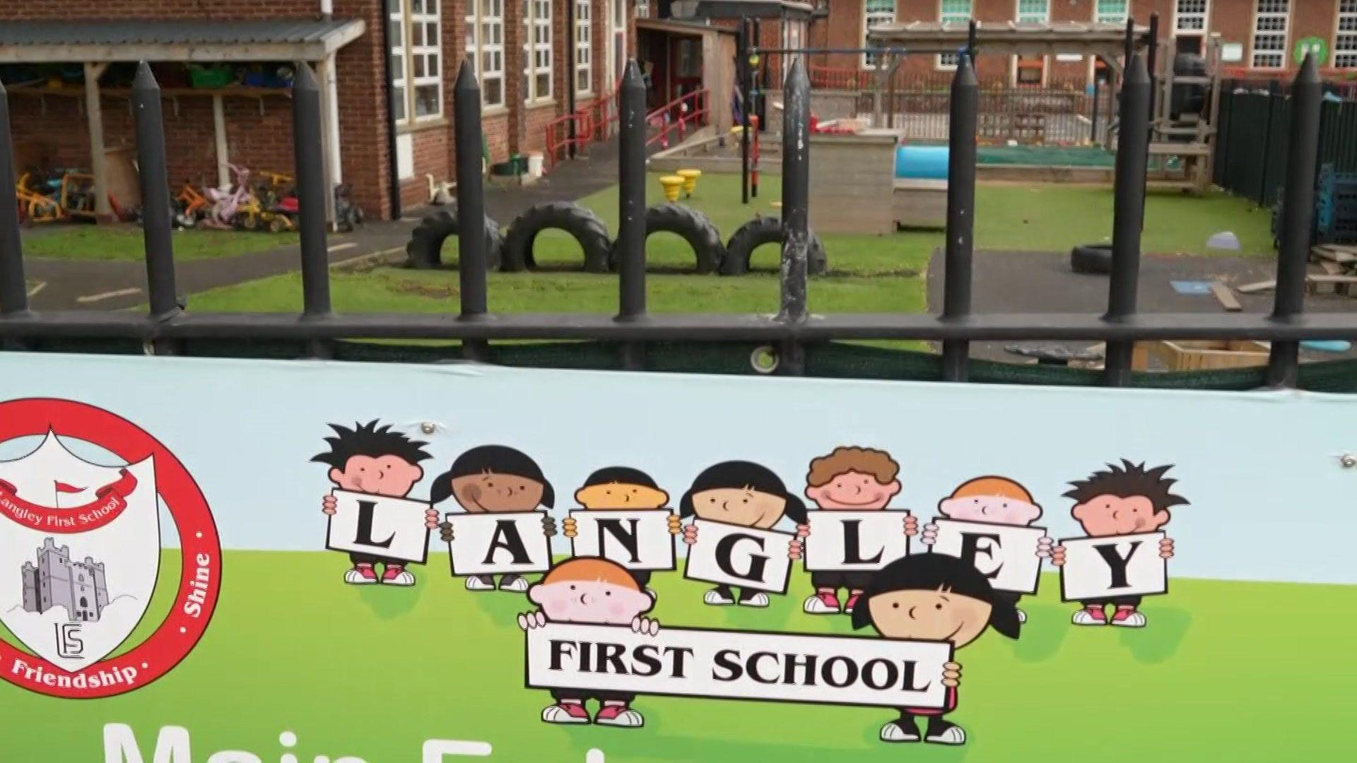 Langley First School which is a red brick building. The playground sits in front of the school with various bits of equipment. In the foreground is a Langley First School sign which is hanging on the black gate.