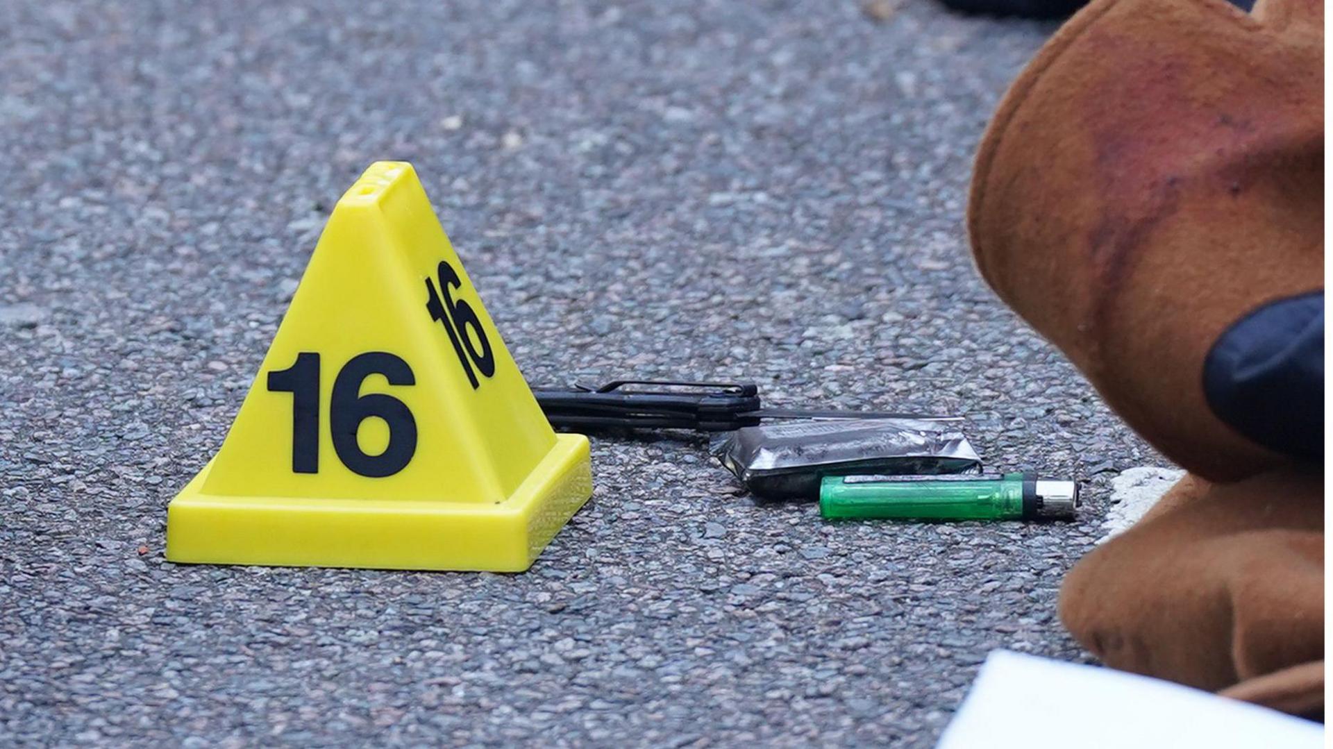 A knife at the scene of the incident with a yellow marker saying 16 on it. There is also a green plastic cigarette lighter on the ground.