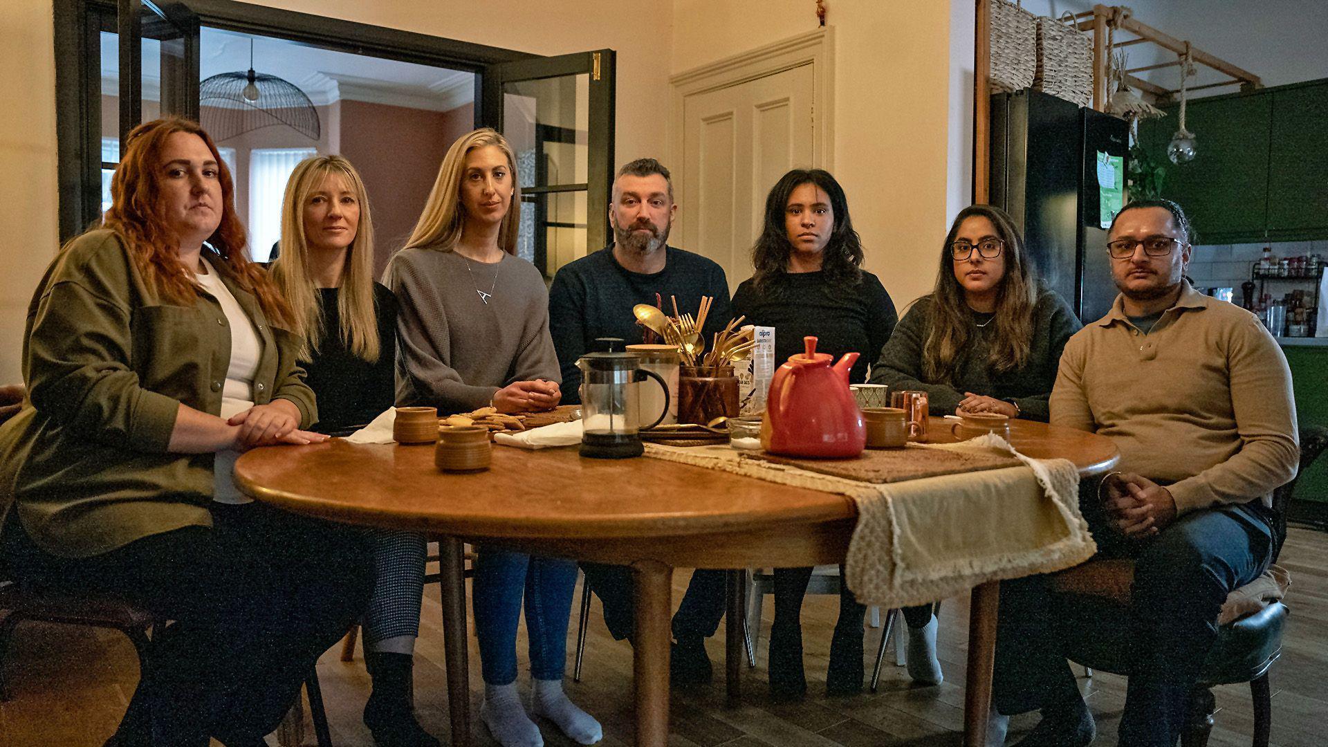 Seven parents - five women and two men - pictured round a wooden dining table looking at the camera with serious expressions on their faces. They include Fiona and Dan, plus Amarjit and Mandip. There is a red teapot, an empty cafetiere and coffee cups on the table.