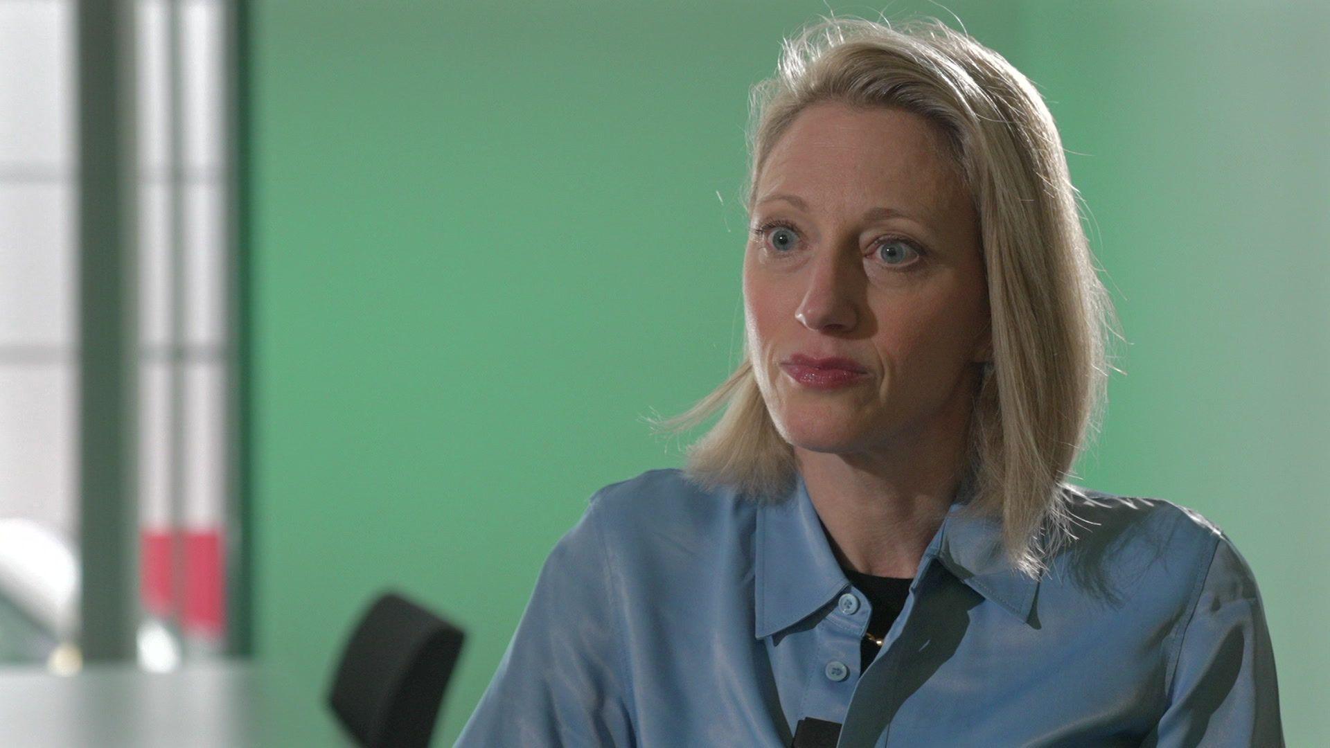 Claire Waxman is seated in an office space, with a green background. She has blonde hair and wears a blue shirt