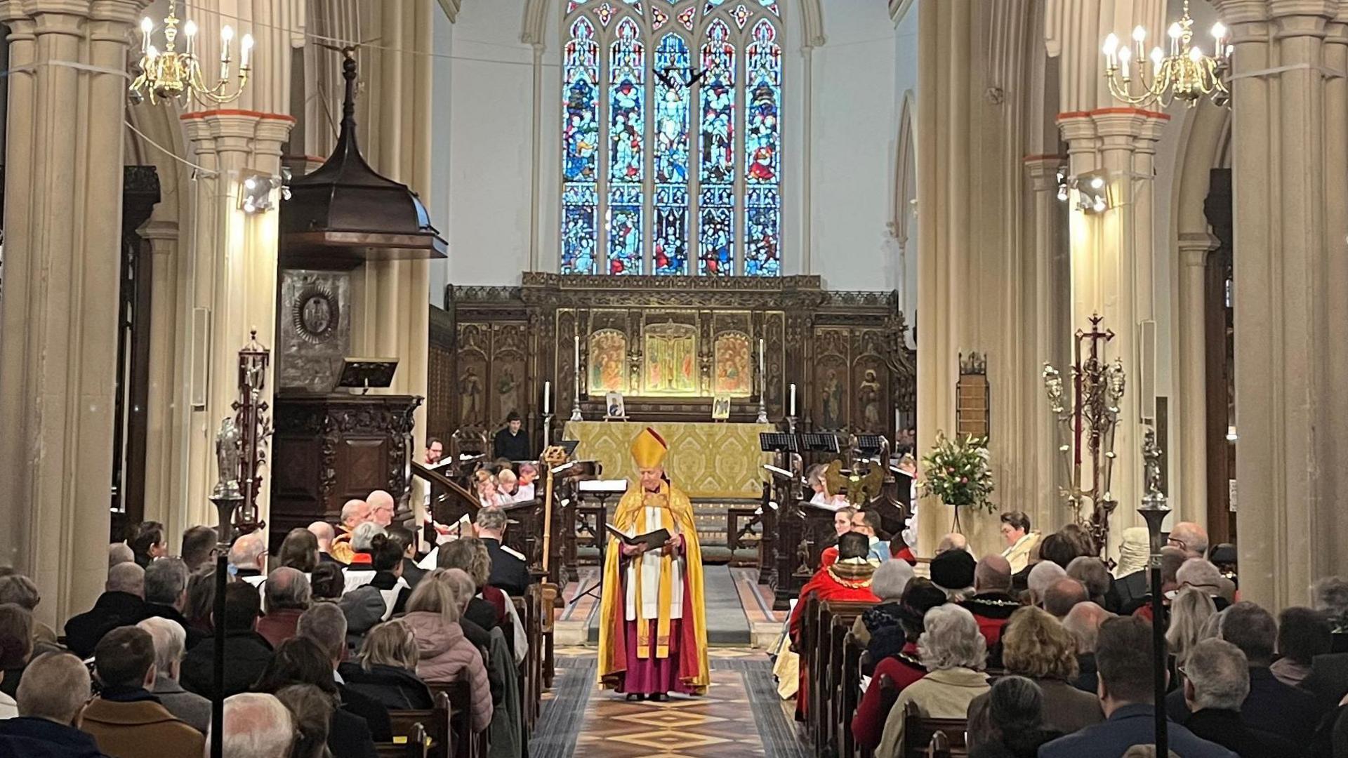 A bishop, in gold coloured robes, stands at the front of a full medieval church congregation. About 30 people can be seen sitting in the pews, facing away from the camera.