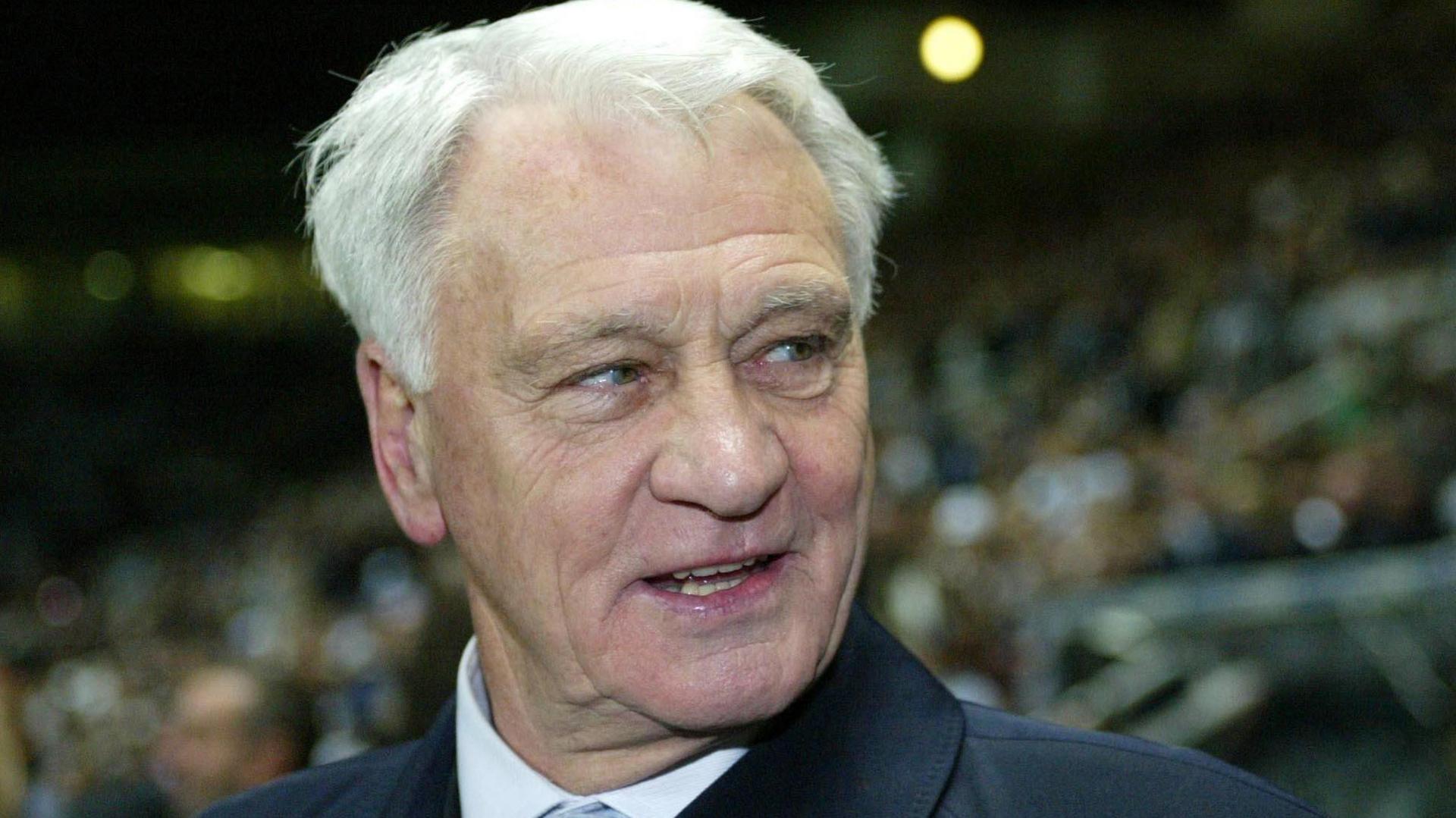 Sir Bobby Robson looking to the side of the camera with a slight smile and his mouth open. He has white, short hair and is wearing a shirt. An out-of-focus football crowd is sat behind him.