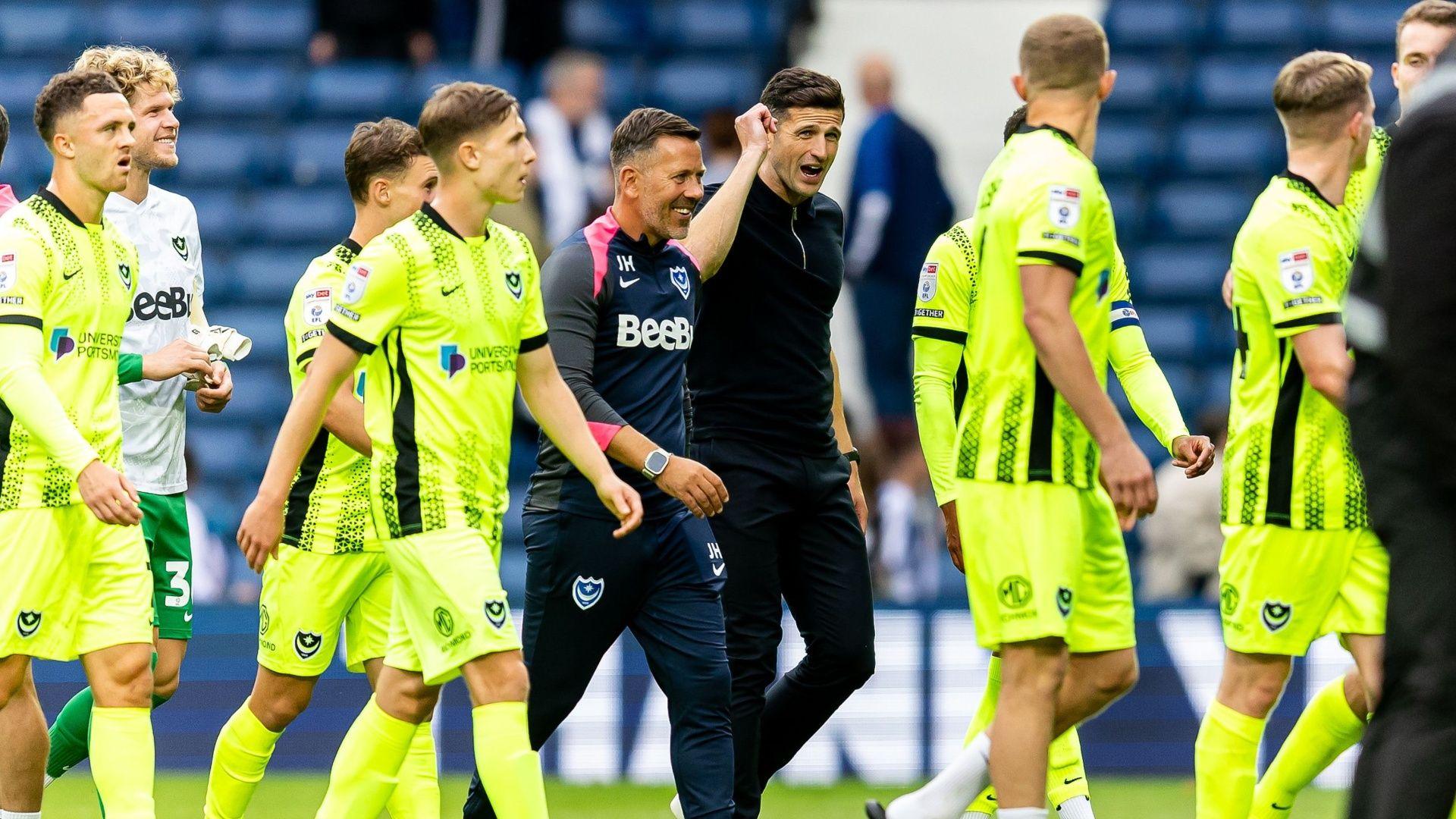 Portsmouth manager John Mousinho walks on the pitch and talks with his players and fellow coach. The players wear a luminous yellow kit and Mousinho wear a black quarter zip top with black trousers