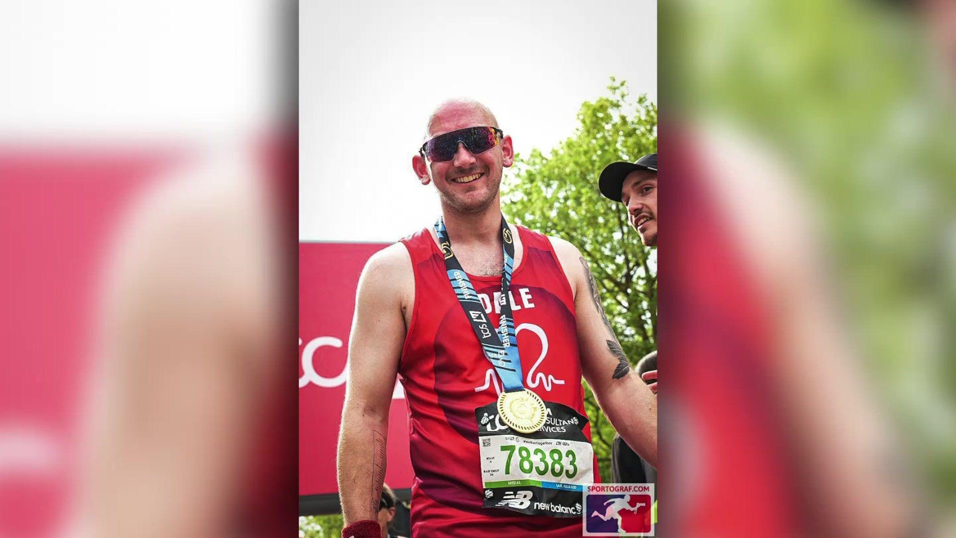 A man at the finish line of a marathon. He is wearing a red vest top, and has a medal round his neck.