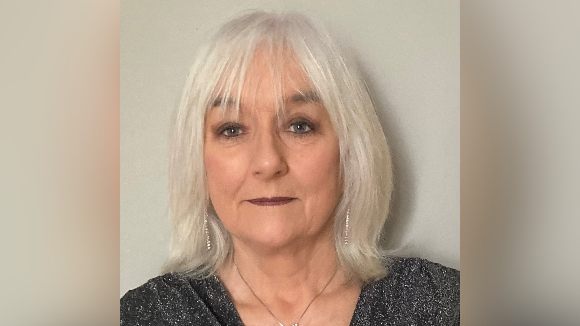 Mary Duggan from the shoulders up, wearing a black top with white speckles on it. She has white shoulder-length hair. She is wearing earrings and a necklace.