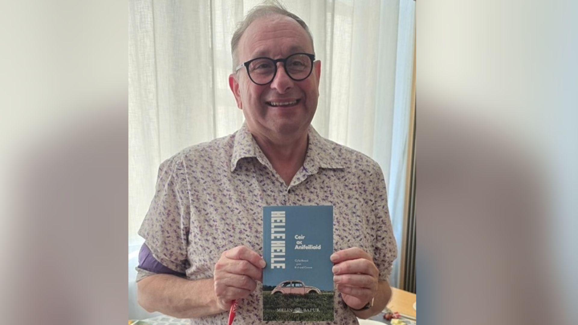 Llun o Richard Crowe yn dal ei gyfieithiad Cymraeg, 'ceir ac anifeilaid'.