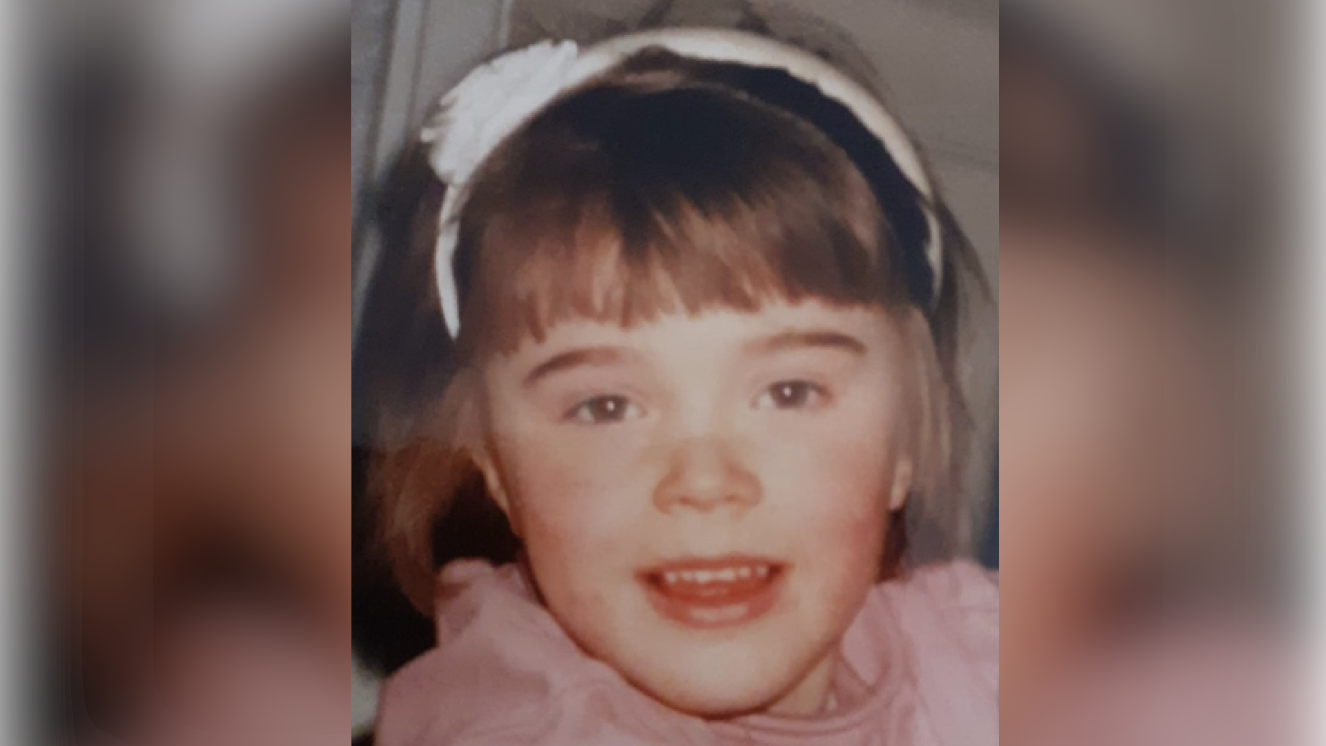 A close-up photo of Emily when she was a child. She is smiling at the camera. She is wearing a white hair band and a pink top.