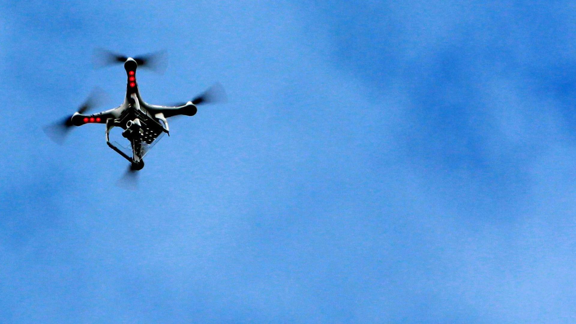 A drone camera with three red anti-collision lights flying in the air. The sky is blue with no clouds.