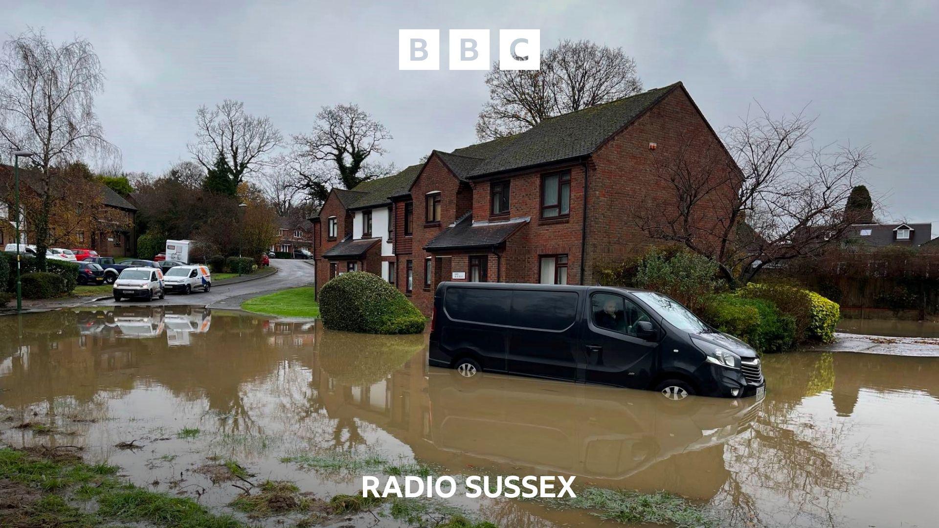 A section of residential road is flooded. A black van is stranded in the flood water, the driver still at the wheel.