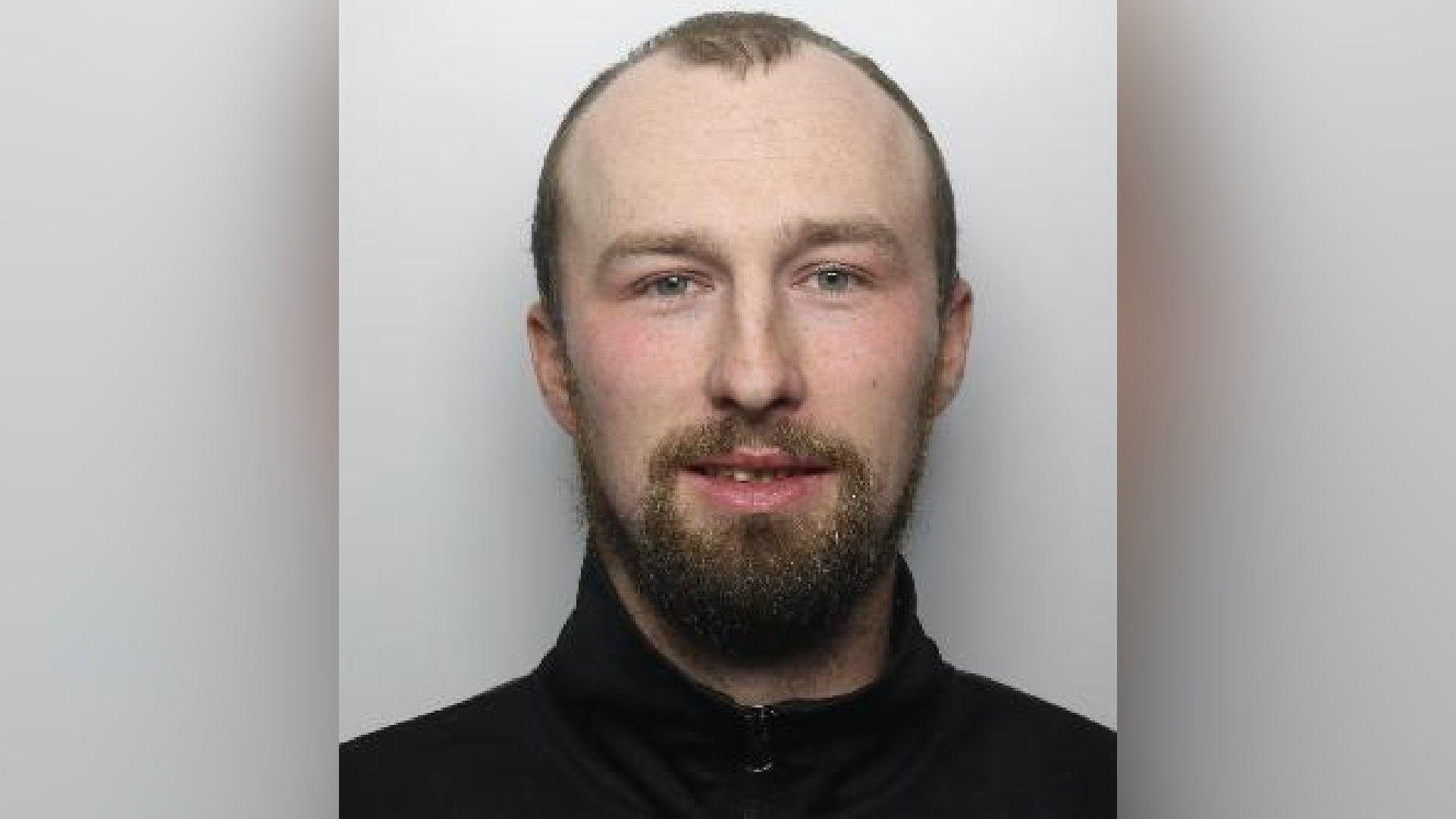 A mugshot of Lane who smiles with his teeth and has a thick goatee. He has a receding hairline with some hair in the centre of his head. 