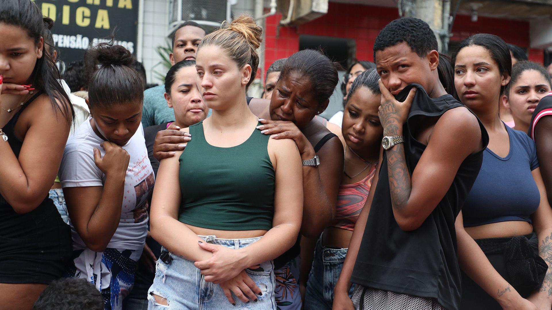 A group of people - many of them women - look at the ground where bodies have been placed. One man is covering his mouth with his T-shirt. A woman is grabbing the shoulders of the woman in front of her and is crying.