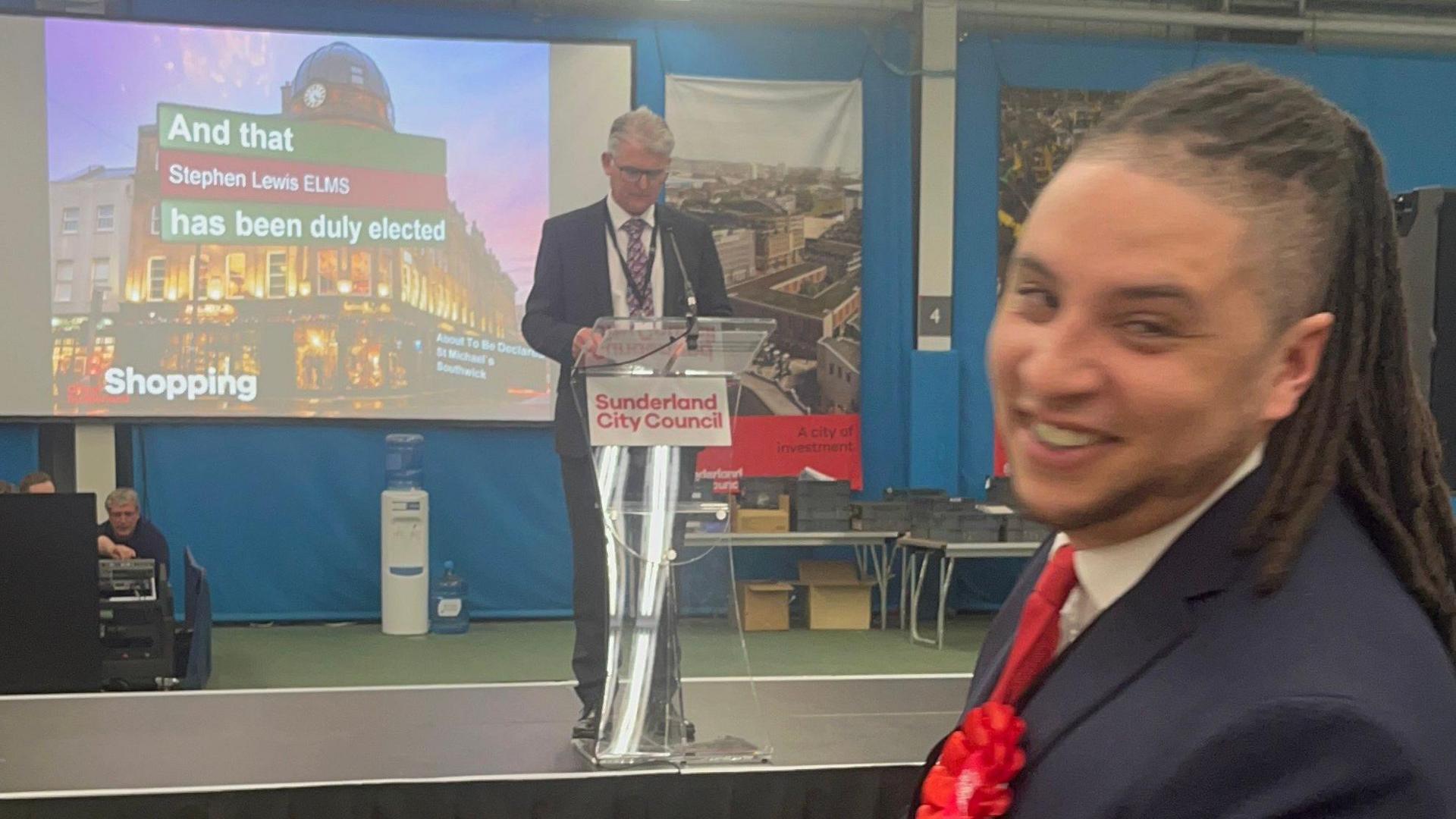 Stephen Lewis Elms, wearing a suit, red tie and a red rosette, is smiling at the camera, as the recorder on the stage confirms he has been elected as councillor. A screen behind the recorder reads "And that Stephen Lewis Elms has been duly elected".