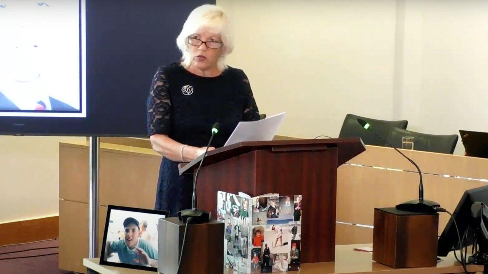 Melanie Leahy speaking at the Lampard Inquiry behind a lectern with photos of her son in front of her on the table