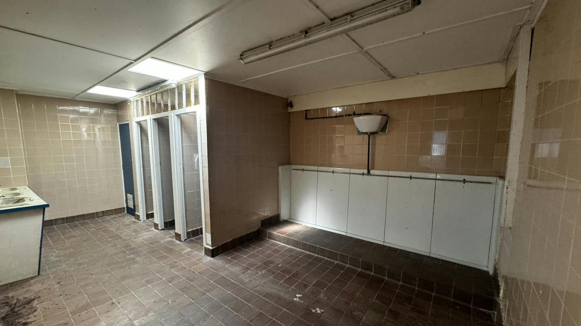 The inside of an old public toilet block. On the left are four cubicles with a large white urinal to the right. The ceiling is white and the floor is brown tiles