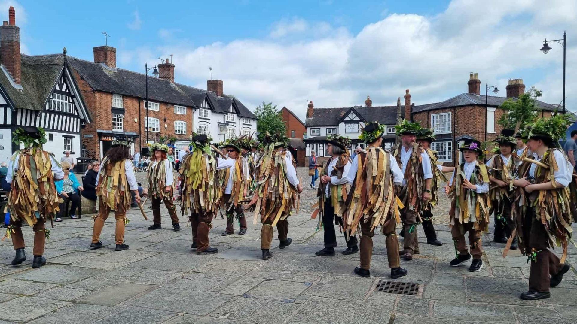 A group of Morris dancers in the centre of Sandbach. They are wearing brown coloured ribbons and green headwear. There are a number of the town's buildings in the background.