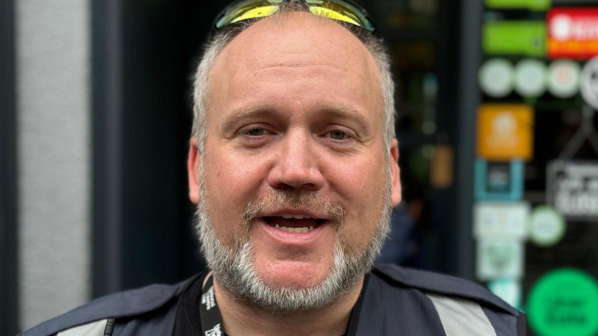 A man with a trimmed beard looks at the camera. He is standing on a street outside a shop front.