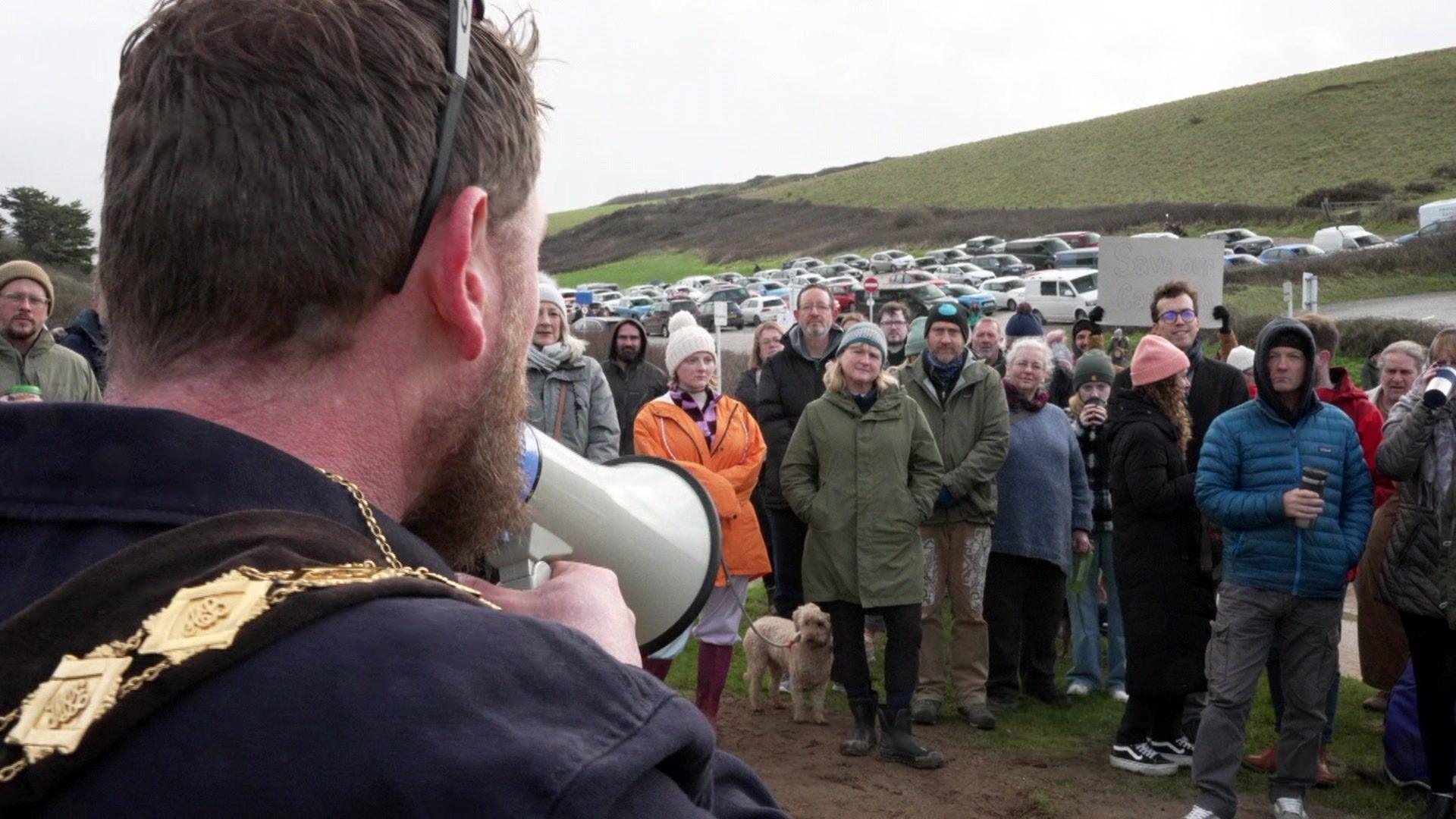 A man holds a megaphone to address a large crowd of people standing on grassy land in warm winter clothing. In the background is a car park and a large slope.