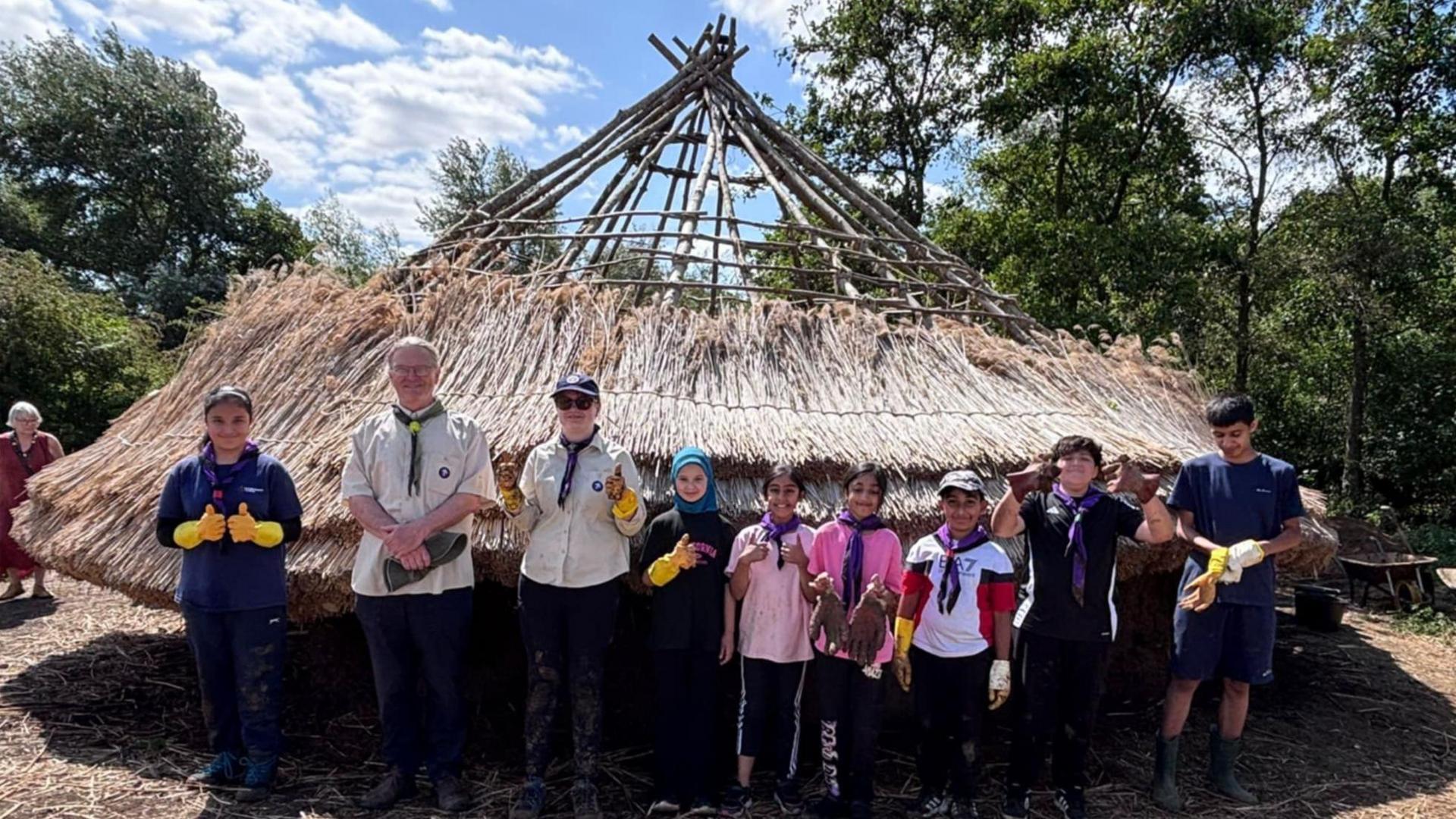 A group of nine people from the Scouts standing outside a partly-built replica Bronze Age roundhouse. There are two adults, a man and a woman, both wearing Scout uniform tops with green scarves and seven children. Many are doing a thumbs up. The roundhouse is a low-lying building with a partly built straw roof. Beyond them are trees.