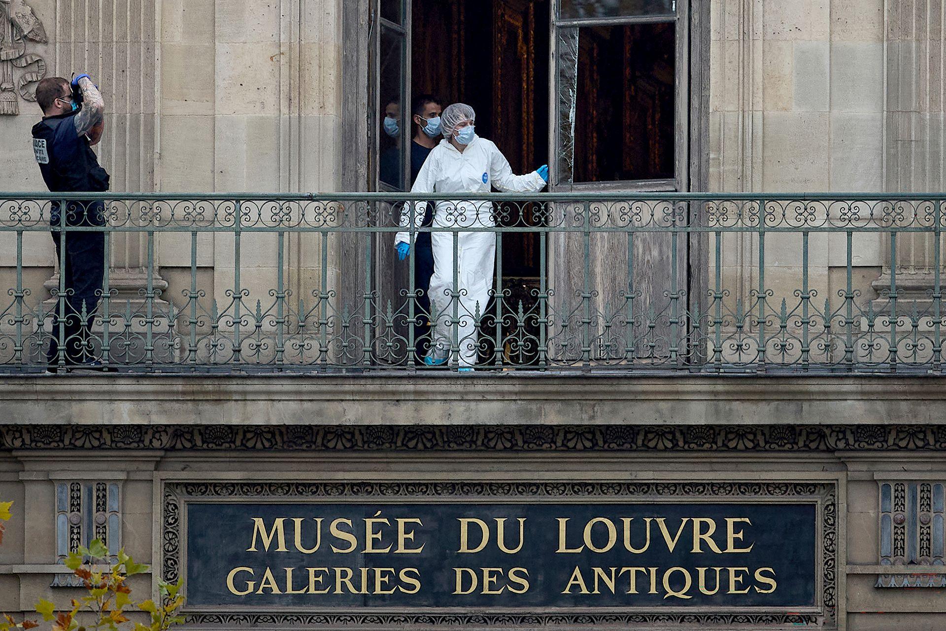 A forensic worker in white overall, gloves and mask exits the balcony door above a sign saying 'Musee du Louvre galeris des antiques' while another officer takes a photo of the scene, in Paris on 19 October.
